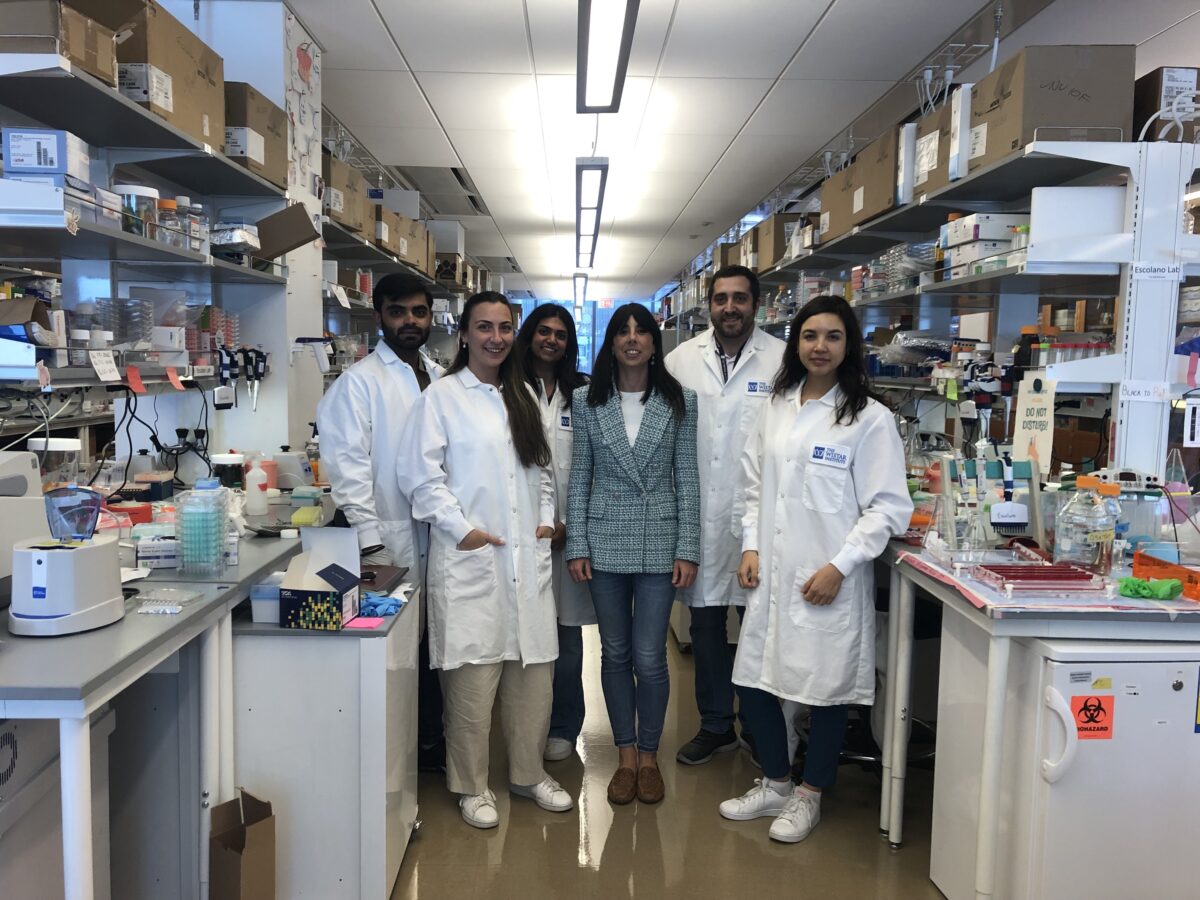 Six people are standing in a laboratory, five wearing white lab coats and one in a blazer. They are surrounded by lab equipment and shelving filled with various supplies.