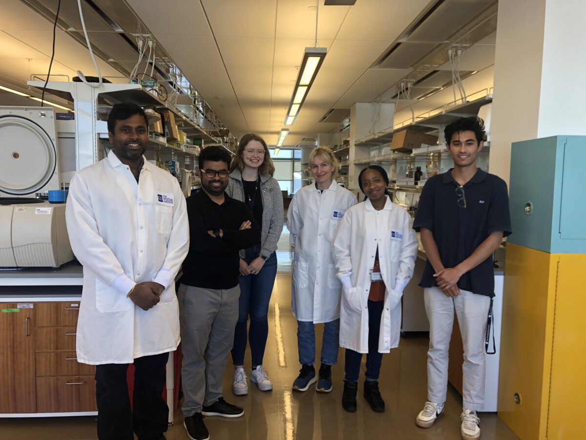 Six people standing in a laboratory, with four wearing lab coats and two in casual attire, posing for a group photo. Laboratory equipment is visible in the background.