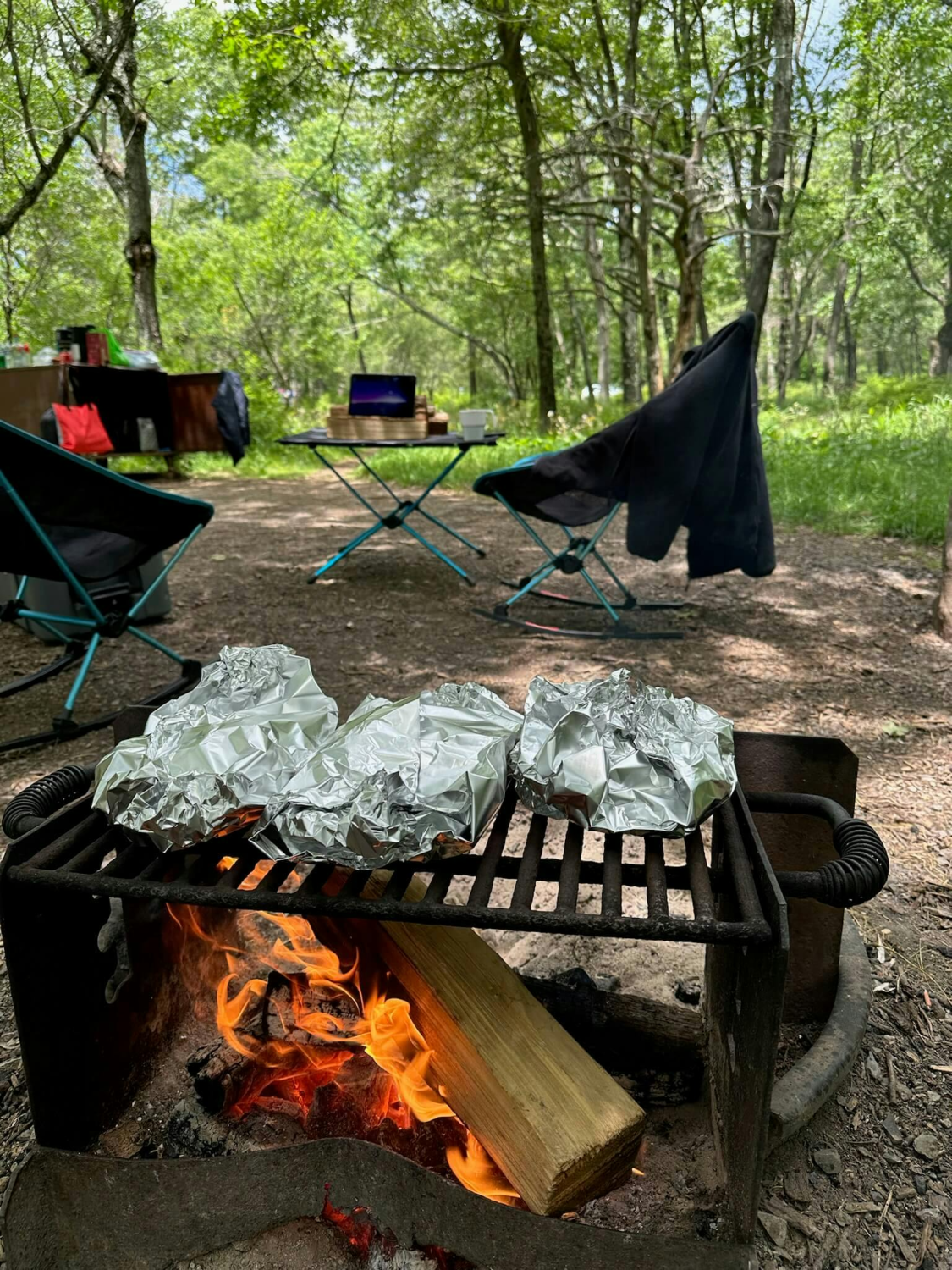 Food grills in foil on black grate over orange fire in campsite.