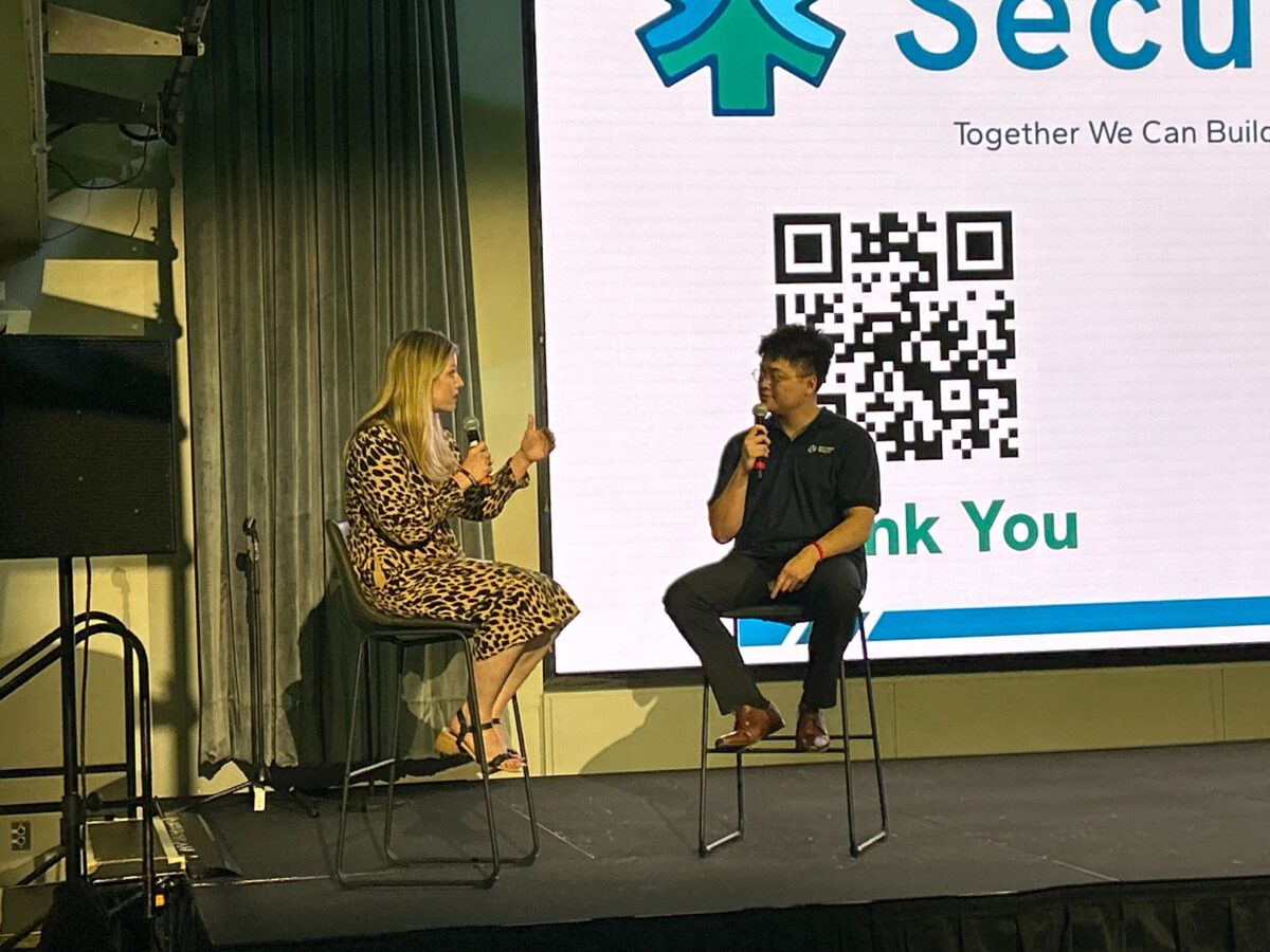 White woman in leopard-print dress speaks to asian man in navy and grey outfit in front of stage with black QR code on screen.