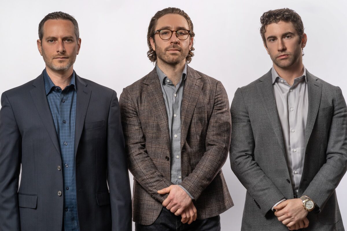 Three white men dressed in business attire stand side by side against a plain white background. The man on the left wears a dark blazer, the middle man wears glasses and a patterned blazer, and the right man wears a light gray blazer.