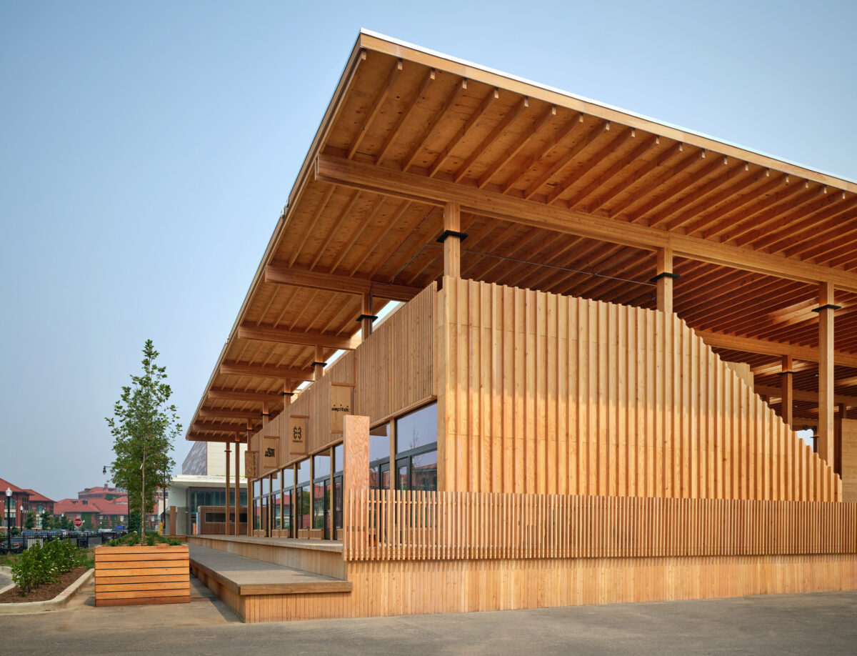 A building with brown slats sits in front of a blue sky and green tree.