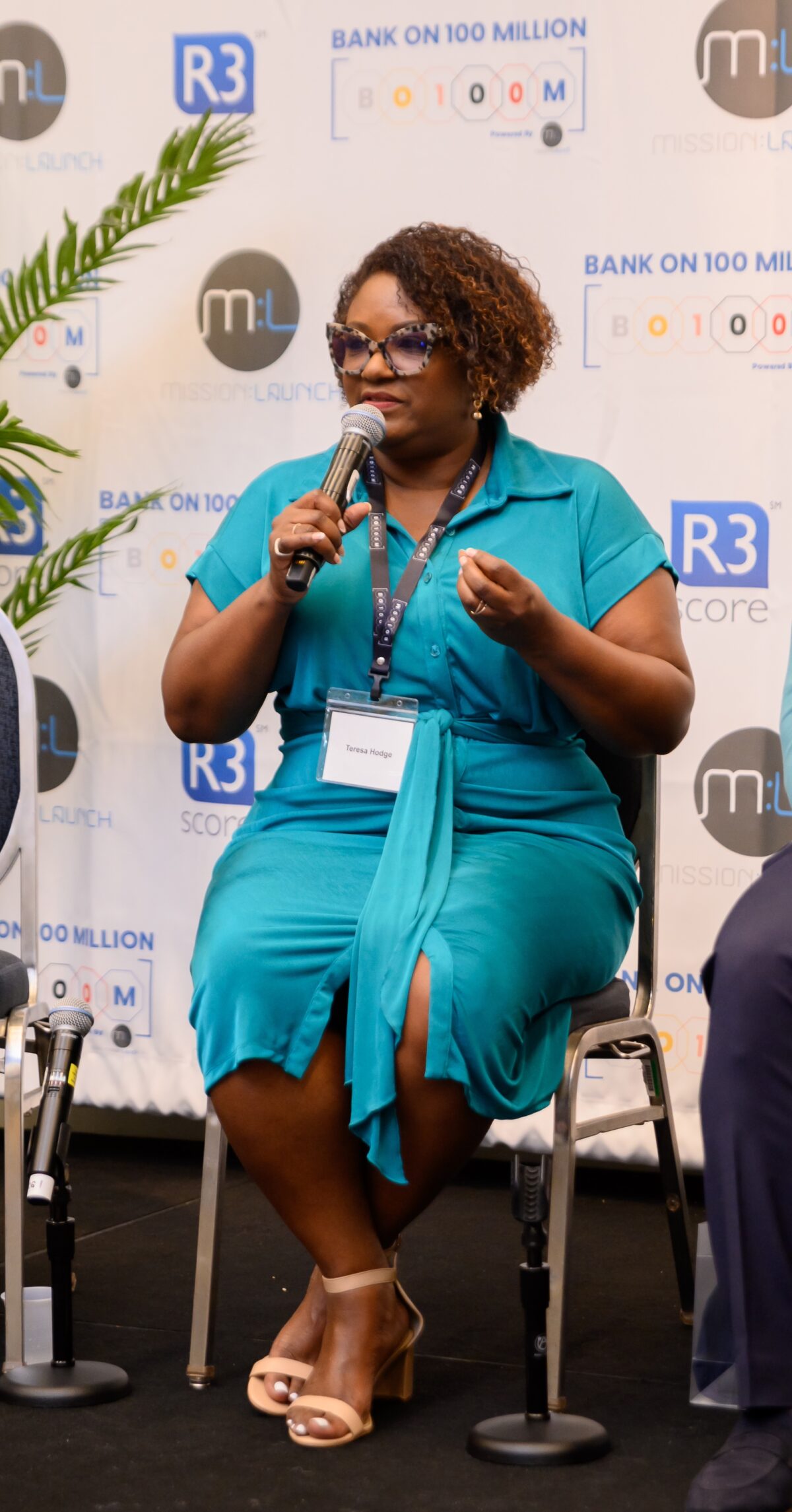 Black woman in a teal dress sits and holds a grey microphone.