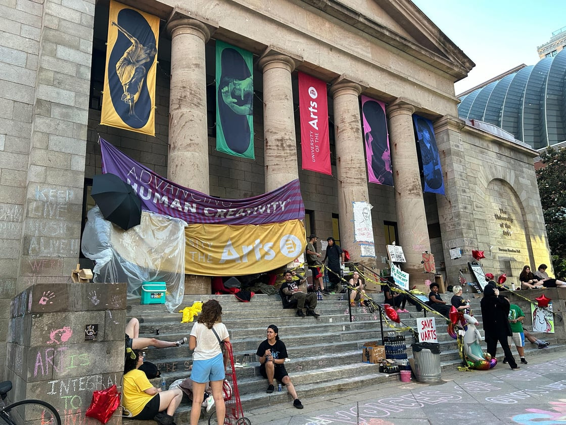 A group of people gather on the steps of a building with banners and signs advocating for arts and humanity. The building features large columns and colorful artwork hanging between them.