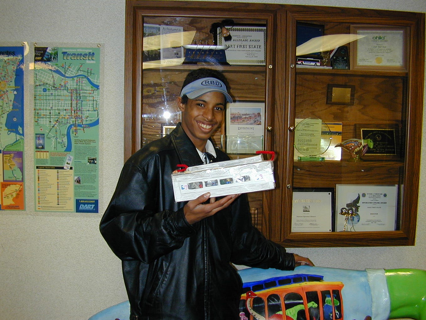 A person wearing a jacket and cap holds a rectangular object with both hands in an office setting, standing beside a colorful display and a wall with maps and certificates.