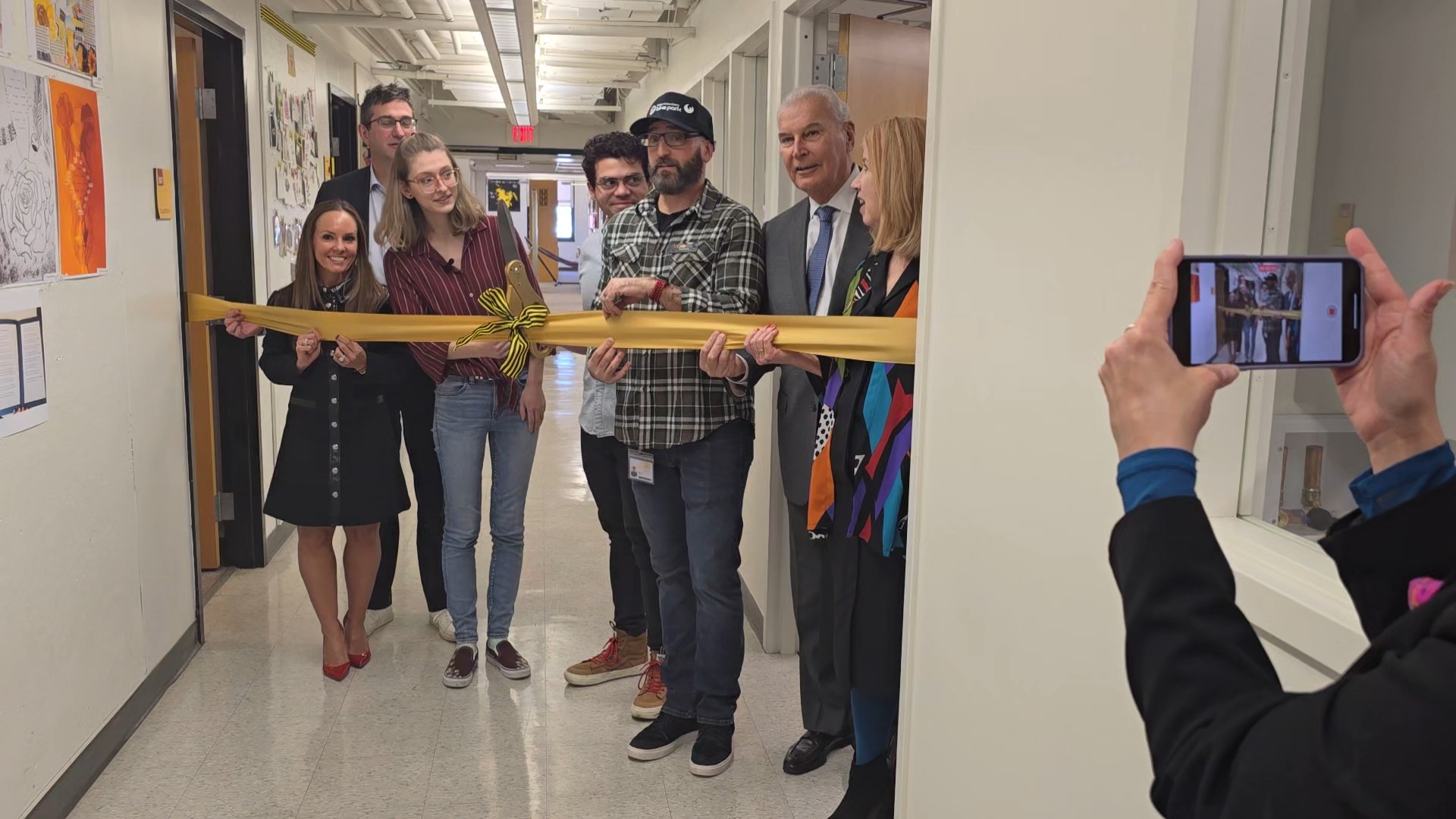 Group of people, including a man and woman holding large scissors, gather in a hallway for a ribbon-cutting ceremony as another person captures the moment on a smartphone.