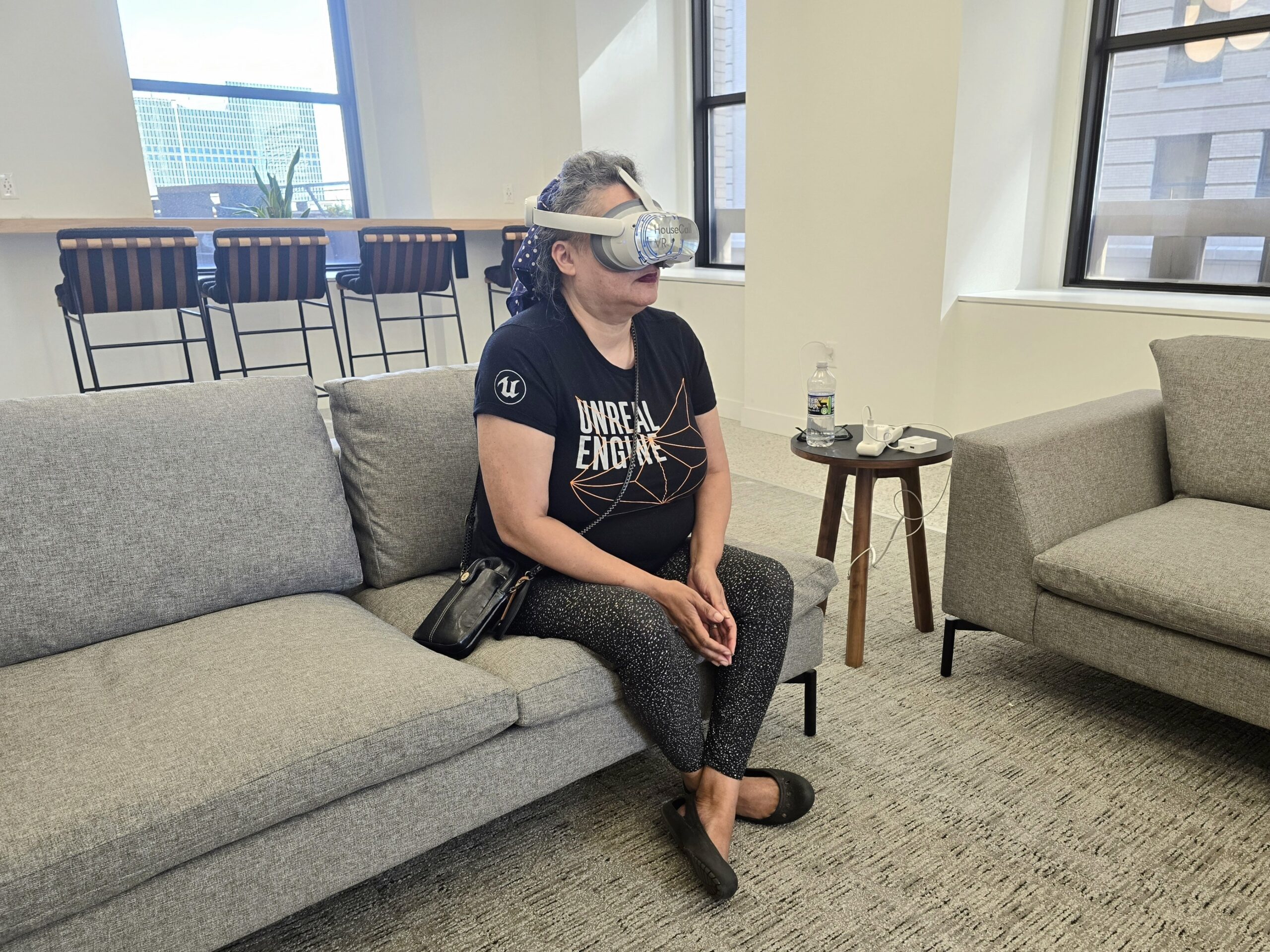 A person sitting on a gray sofa in an office, wearing a virtual reality headset. Several tall windows and a high counter with stools are visible in the background.