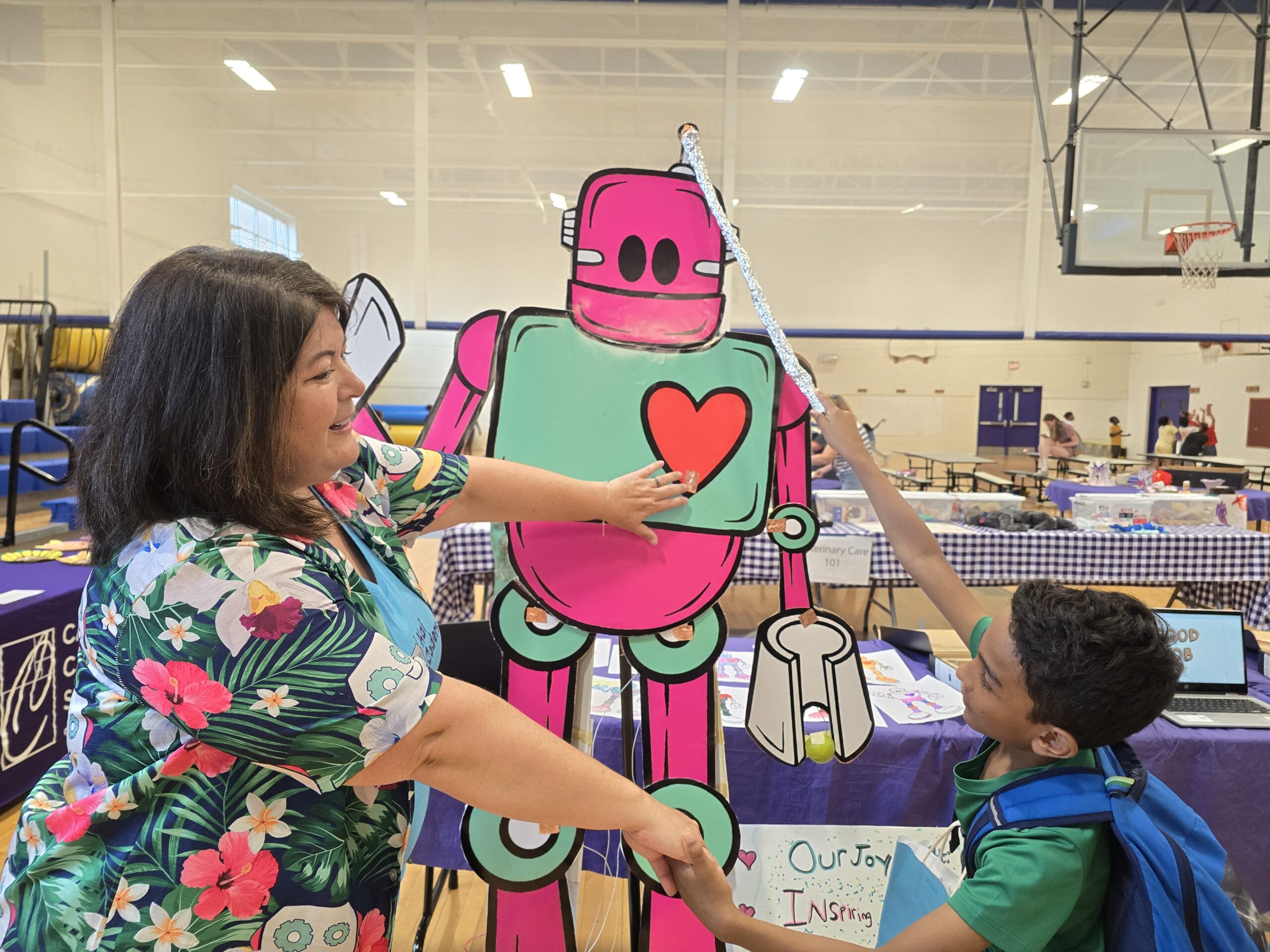 A woman and a child interact with a colorful robot cutout at an indoor event with various booths in the background. The boy points at the robot with a stick while the woman holds its hand.