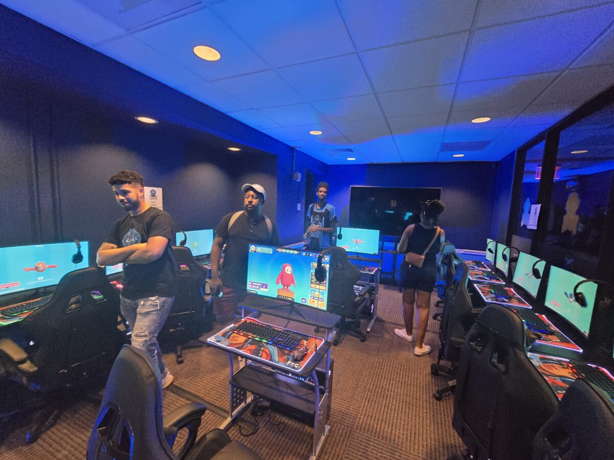 A group of people stand in a dimly lit room filled with multiple computer stations displaying gaming screens under blue lighting.