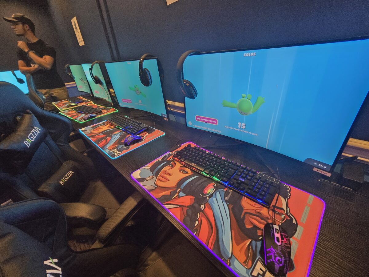 A row of gaming stations with large monitors, headsets, and customized mouse pads, each displaying the Fall Guys game. A person stands in the background next to the equipment.