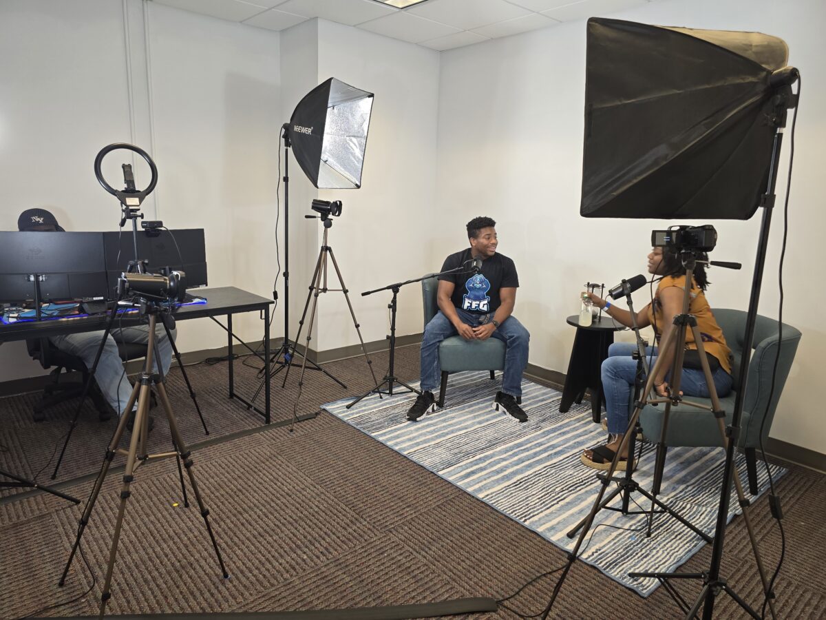 Two people sit in armchairs facing each other in a recording studio setup, with cameras, lights, and audio equipment surrounding them.