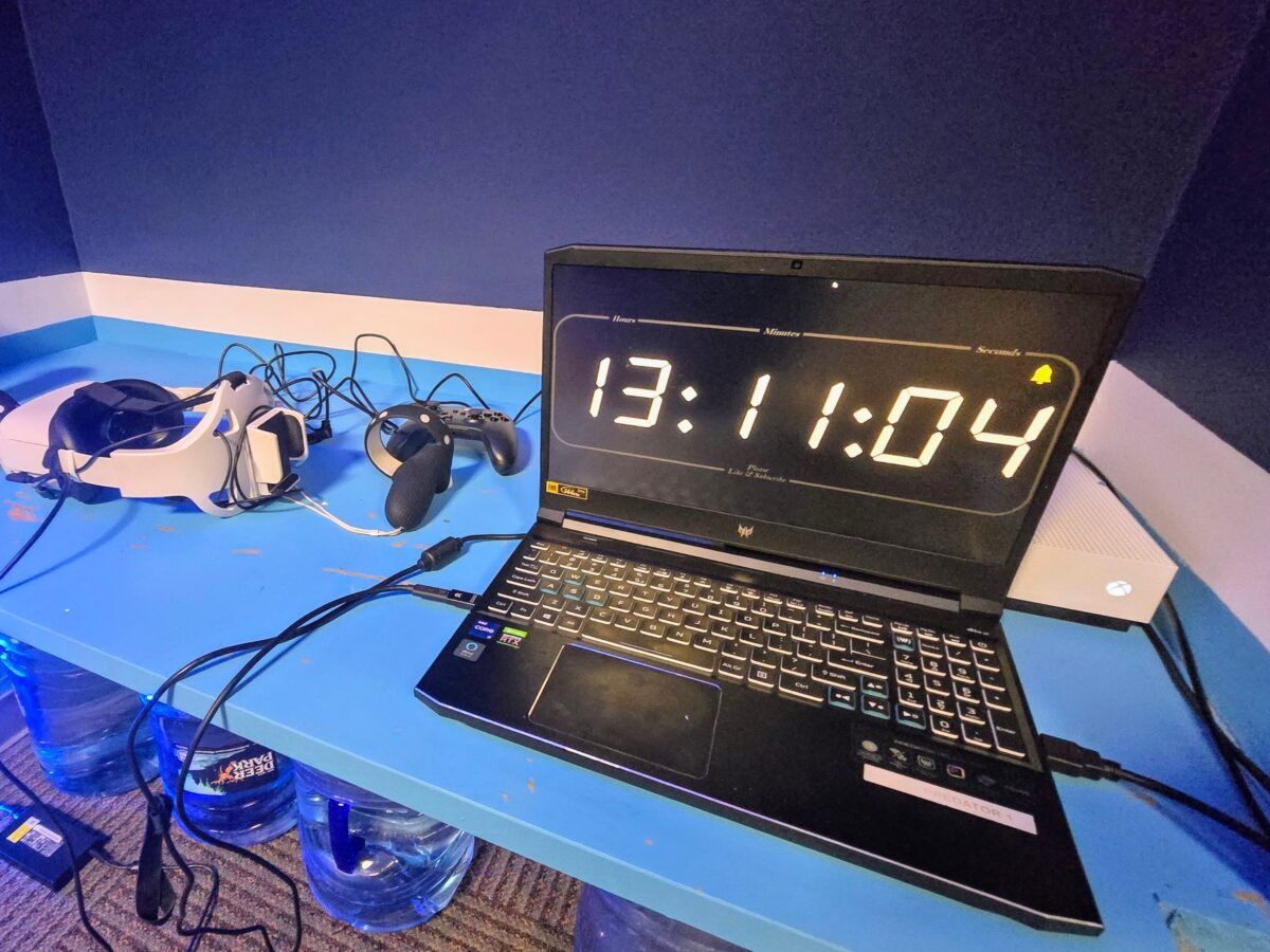 A laptop with a large digital clock on its screen next to a VR headset, a game controller, and an Xbox console sit on a blue desk.