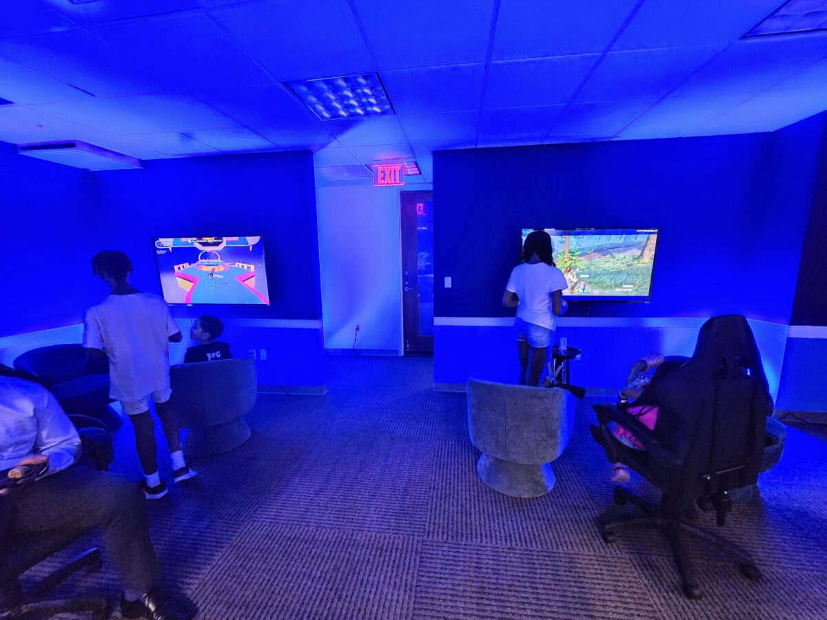 People are playing video games at consoles in a room with blue lighting. Two individuals are standing, while others are seated on chairs facing large screens.