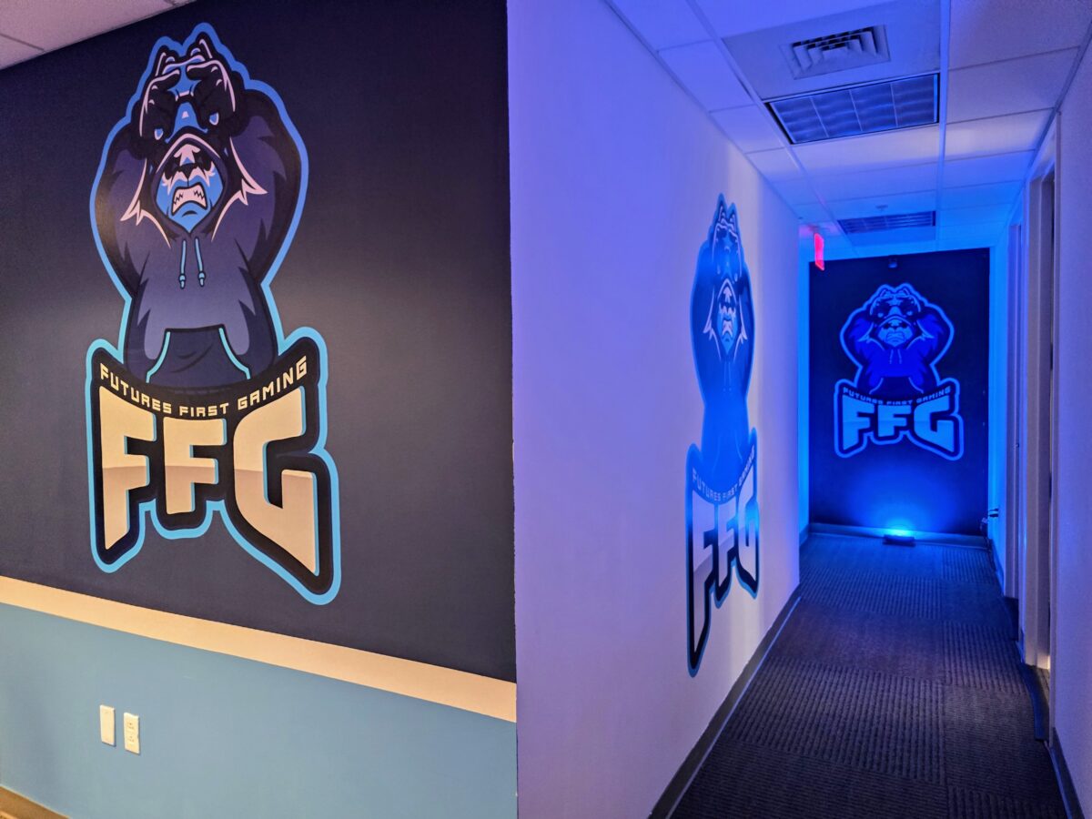 A hallway with logos of a bear wearing a hoodie labeled "Future First Gaming" and "FFG" on the walls, illuminated by blue lighting.