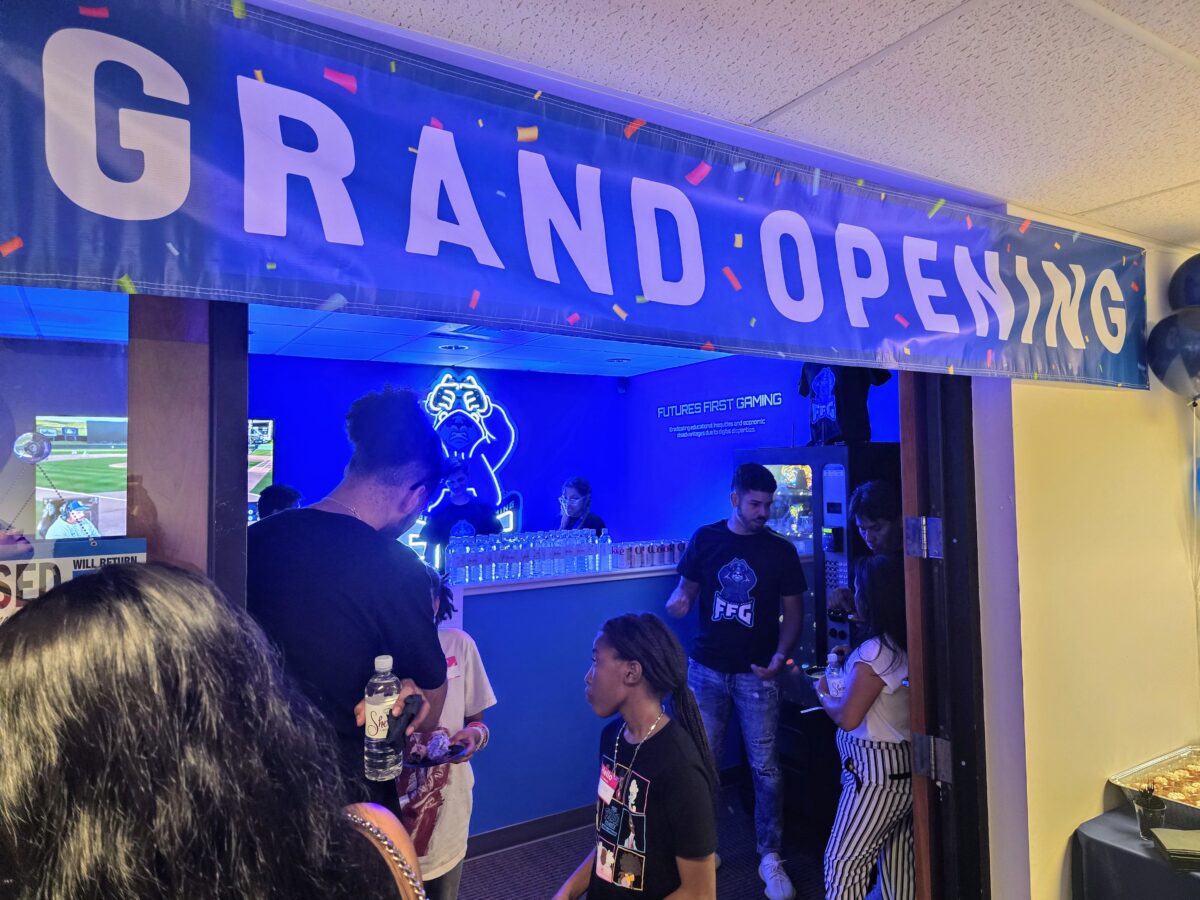 People gathered at the entrance of a room with a “Grand Opening” banner above; inside, a neon sign and video game screens are visible.