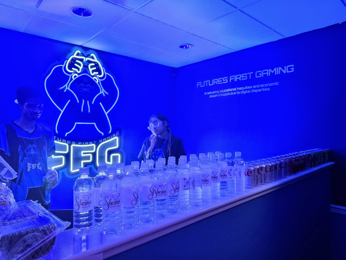 A person stands behind a counter with assorted drinks in a brightly lit, blue-themed room featuring a neon Futures First Gaming logo and a wall sign about empowering through digital capabilities.