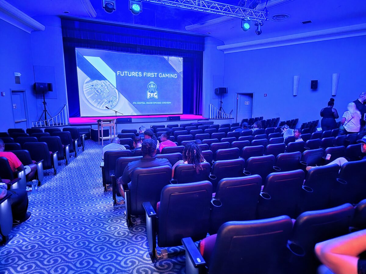 A sparsely filled auditorium showing a screen with the title "Futures First Gaming" before a presentation. Some attendees are seated while others stand or walk in the background.