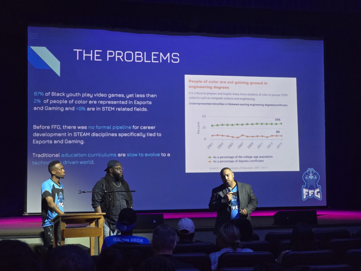 Three speakers present on stage in front of an audience with a projector screen displaying information about the lack of diversity in STEM fields and gaming, accompanied by a graph and text.