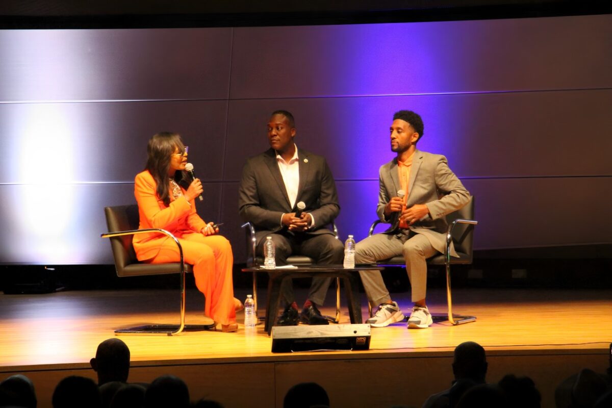 Black woman in orange pantsuit talks to two Black men in dark-colored suits on brown stage.