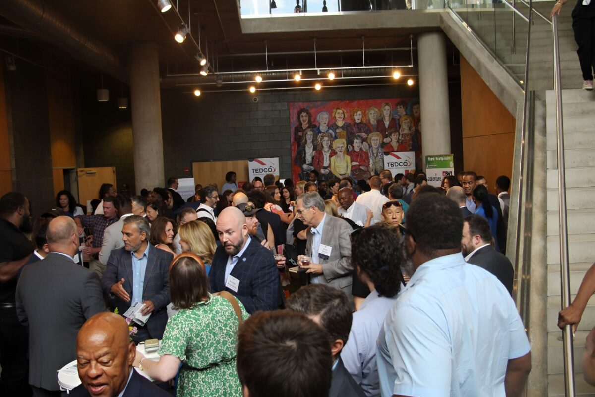 Crowd stands in front of TEDCO signage and mural woth women in grey-walled room