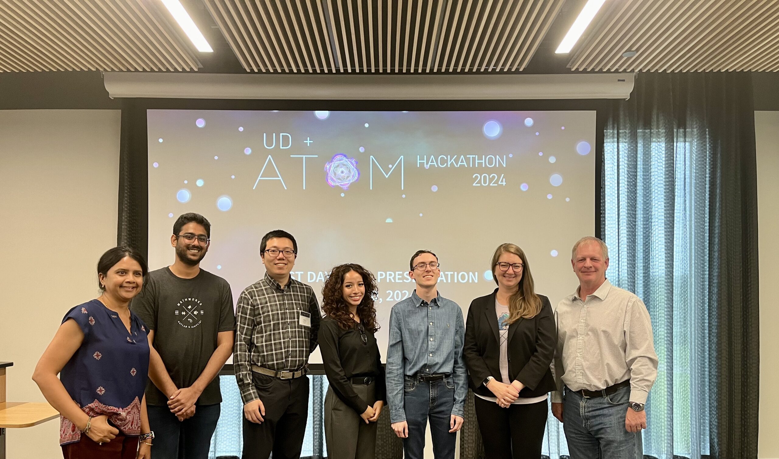 A group of seven people standing in front of a screen displaying "UD + ATOM Hackathon 2024." They are smiling and posing for the photo in a conference room.