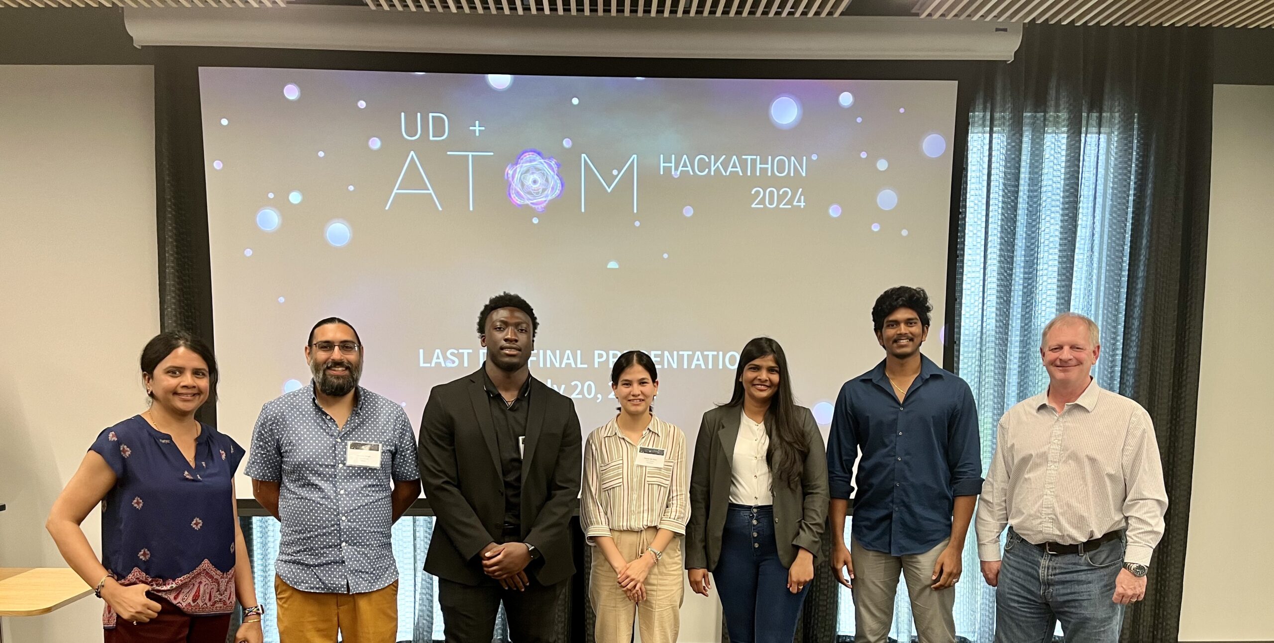 Seven people standing in a line indoors, smiling at the camera. Behind them, a screen displays "UD + ATOM Hackathon 2024" and "Last Final Presentation.