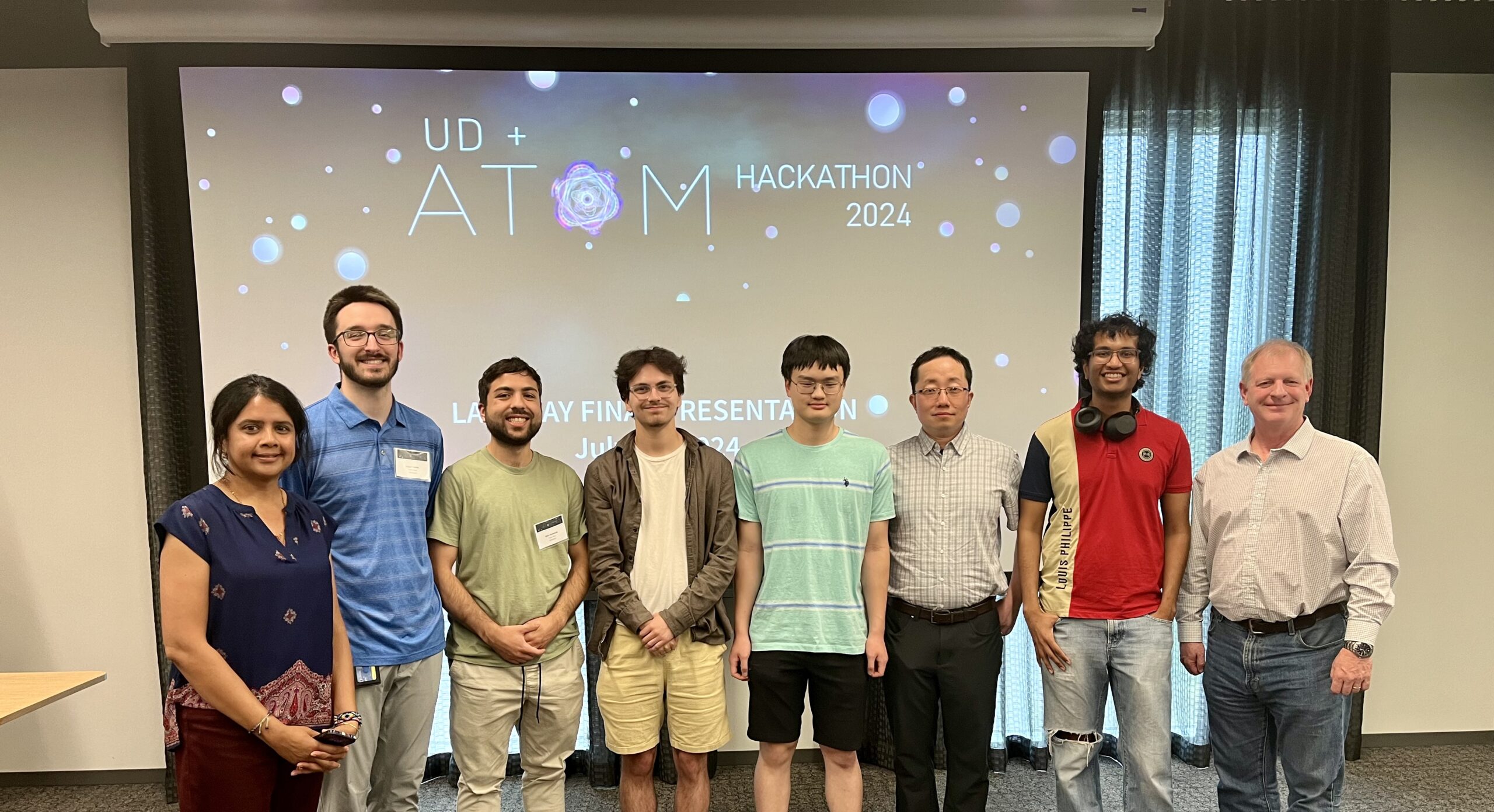 A group of eight people stands in front of a screen displaying "UD + ATOM Hackathon 2024." They are smiling and posing for a photo.