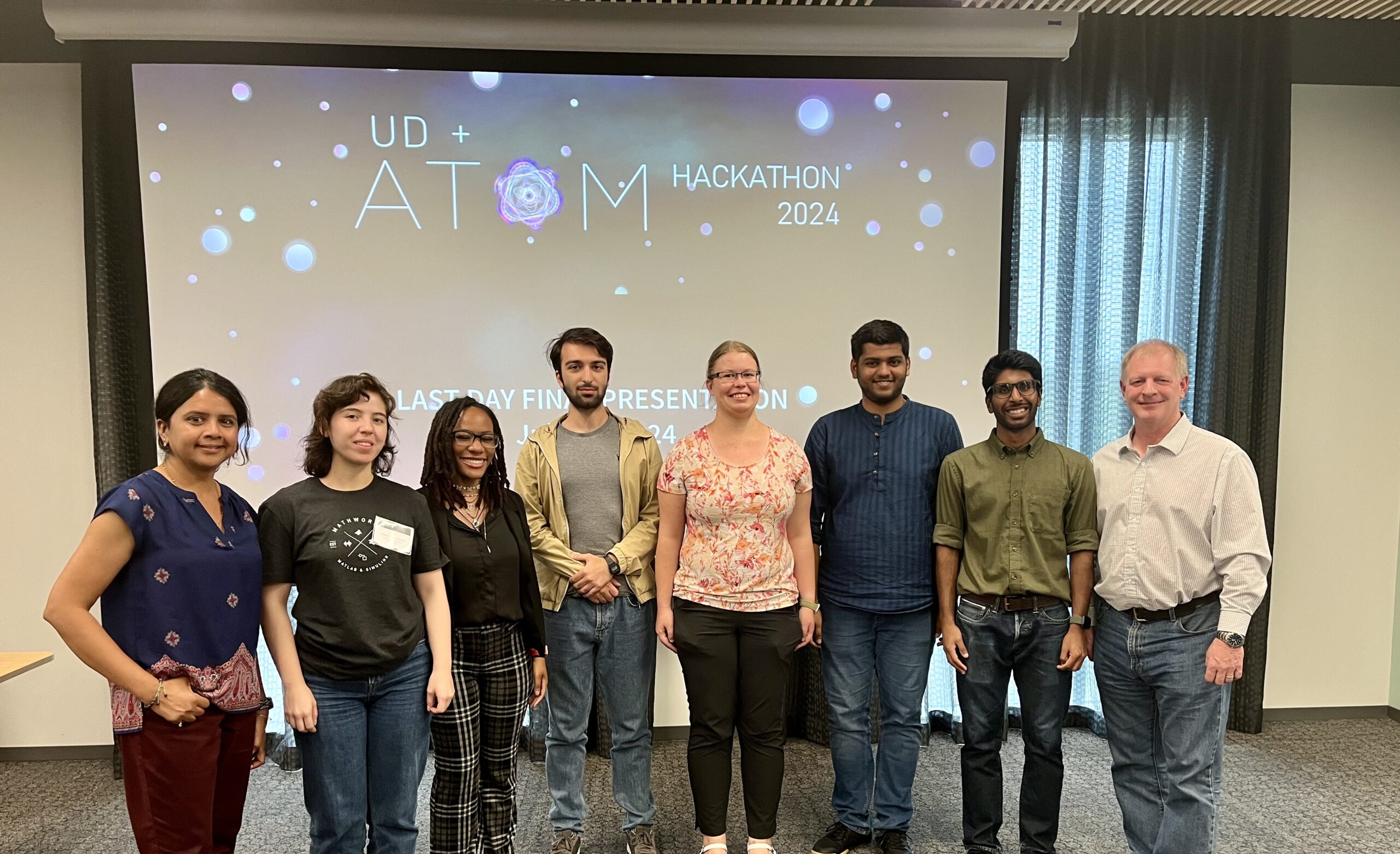 A group of eight people stand in front of a screen displaying "UD + Atom Hackathon 2024." They are posing for a photo and smiling.