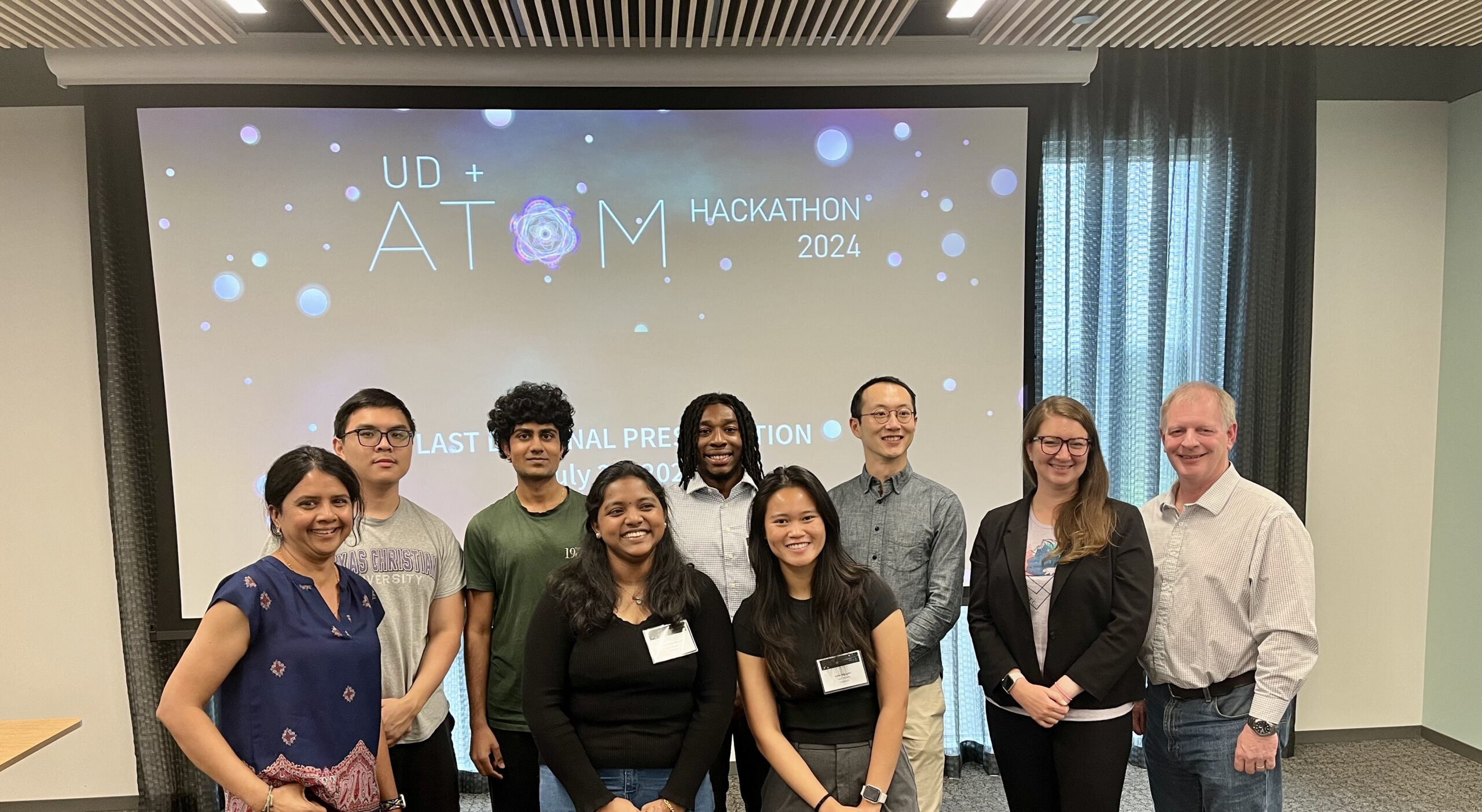 A group of nine people standing in front of a projector screen displaying "UD + ATOM Hackathon 2024". Some are smiling, and they appear to be in a conference room.