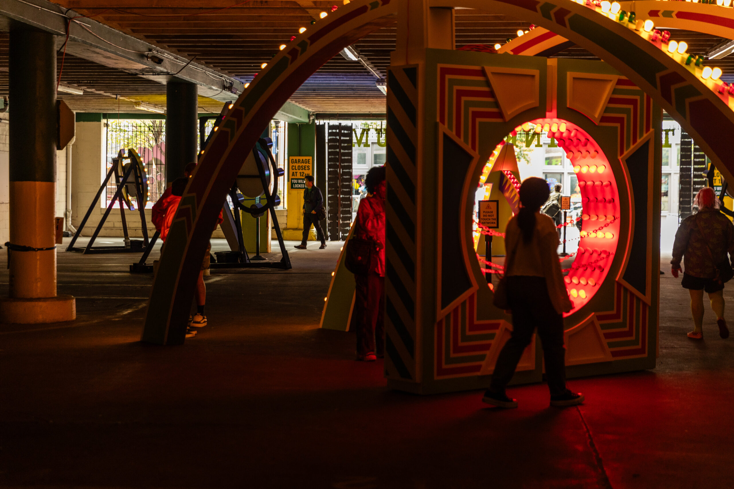 People walk through an illuminated circular installation in a dimly lit, colorful indoor space with various art structures.