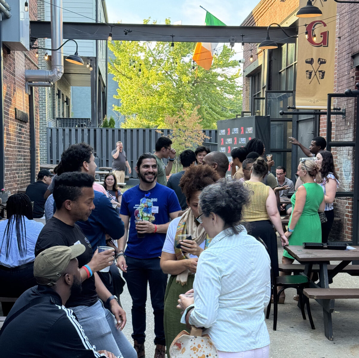 A group of people socializing outdoors in an alleyway with benches and tables, some holding drinks.