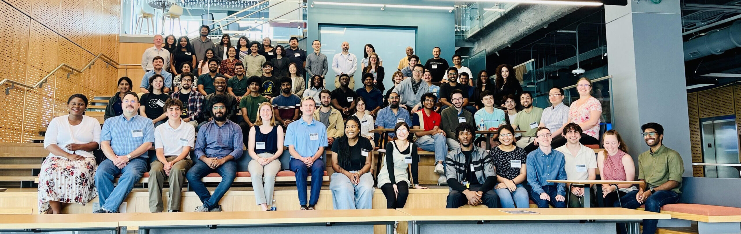 A large group of people of diverse ages and backgrounds is seated and standing in an open, modern indoor area with wooden seating and exposed ceiling pipes. They appear to be posing for a group photo.