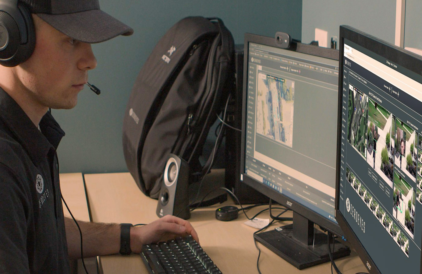 A white person wearing a headset and a cap is working at a desk with two computer monitors displaying surveillance footage. A backpack and a speaker are seen on the desk.