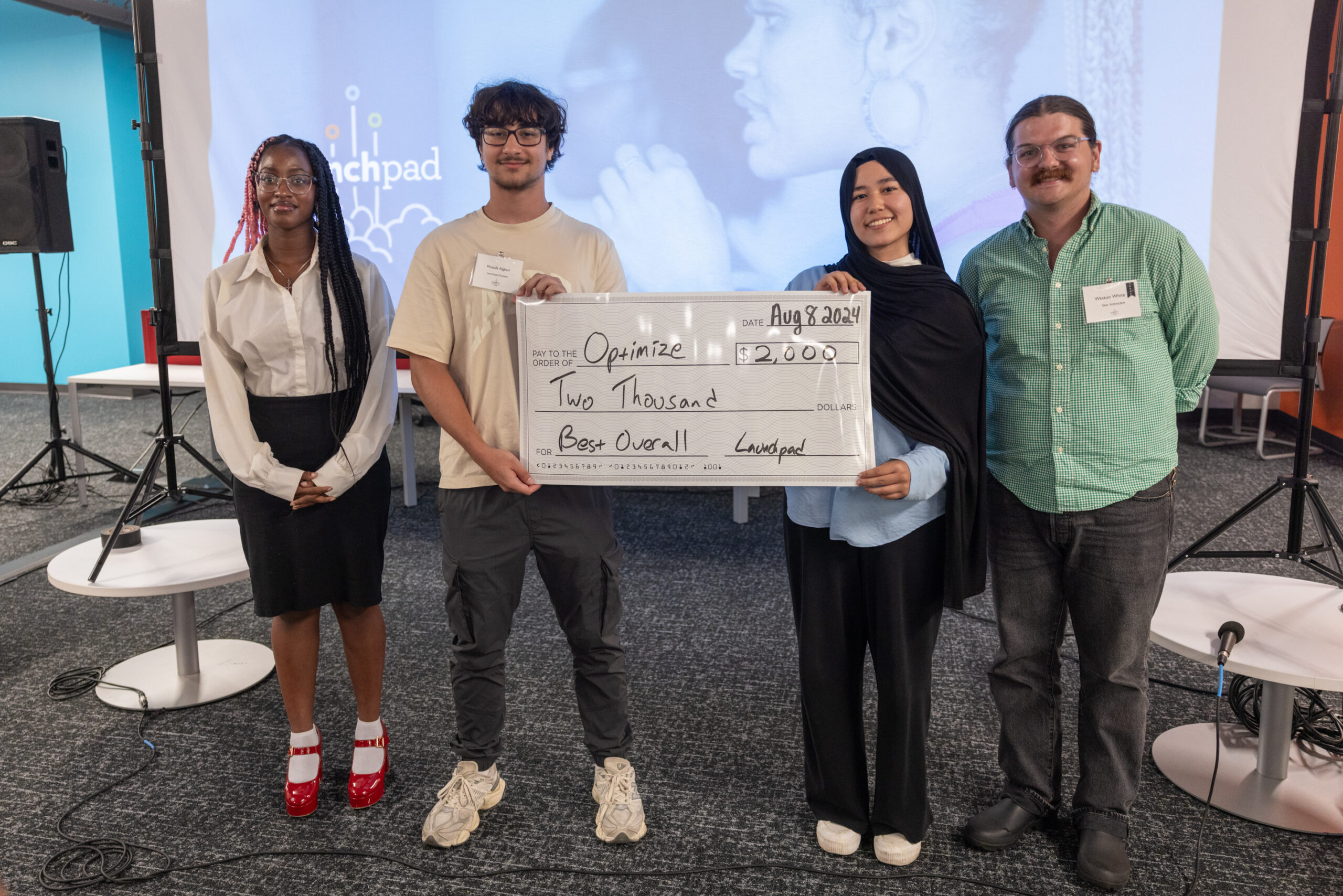 Four people stand together holding a large check for $2,000 with "Best Overall" written on it at a presentation event.