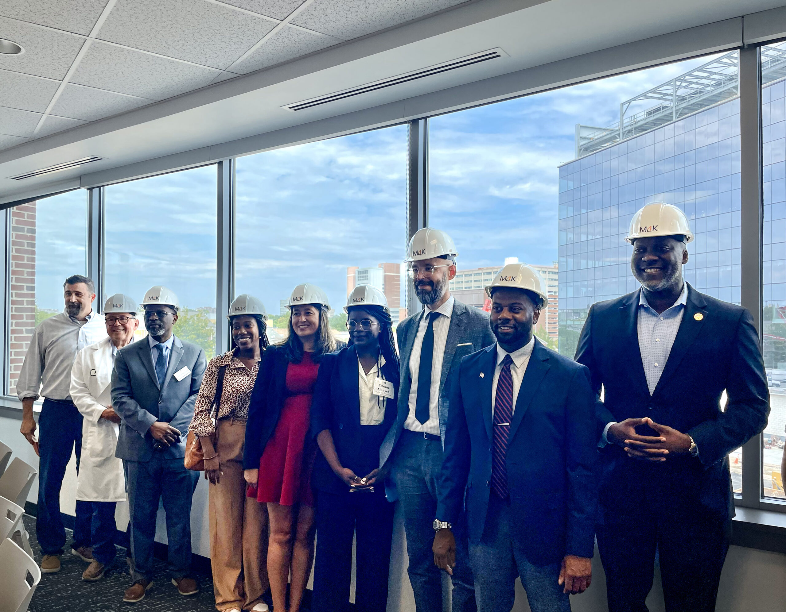 A group of nine people stands in a line indoors, all wearing white hard hats. They are in a room with large windows overlooking urban buildings.