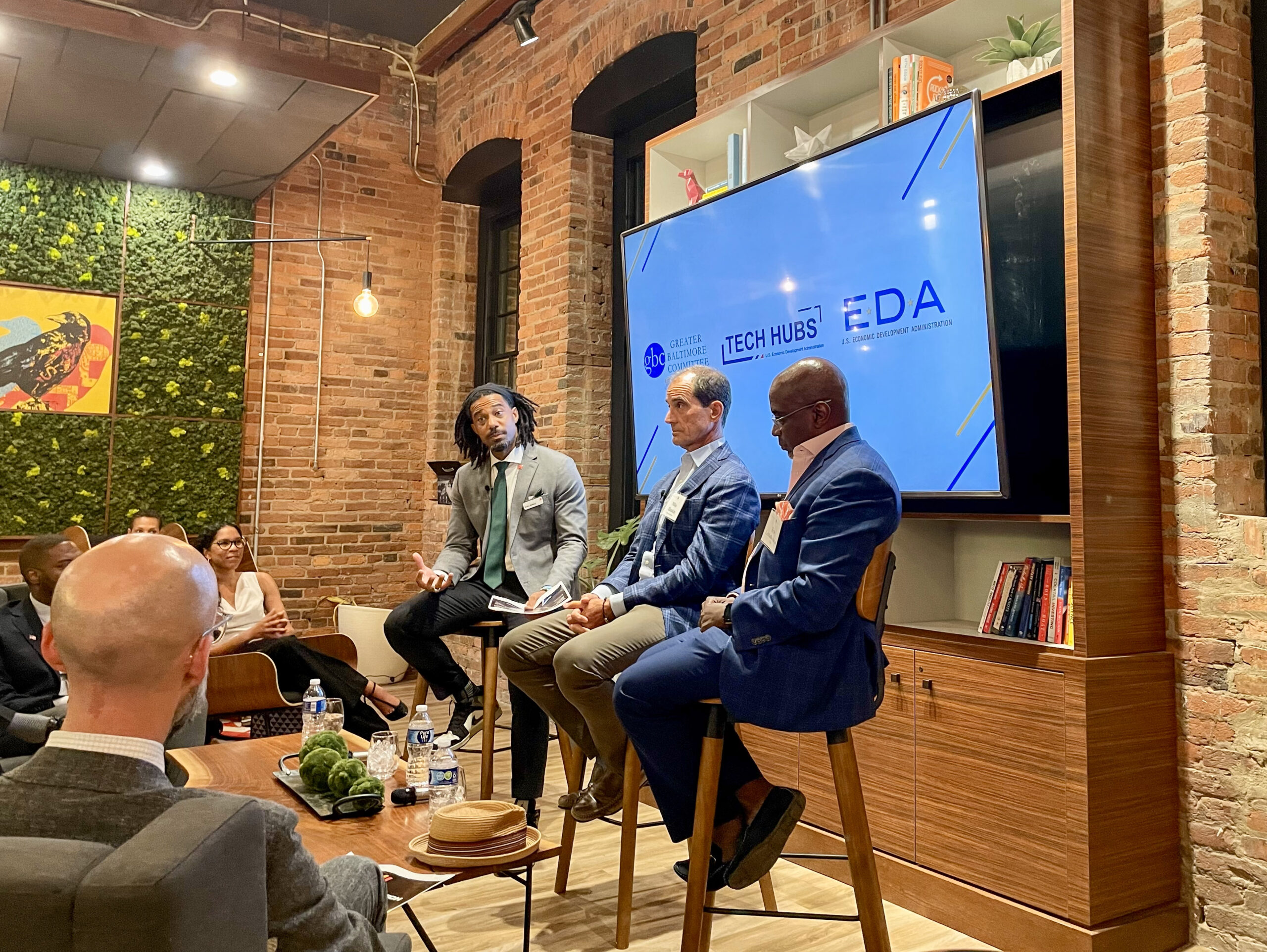 Three men seated on stools in front of a large screen display during a discussion panel in a modern, brick-walled room with an audience watching.