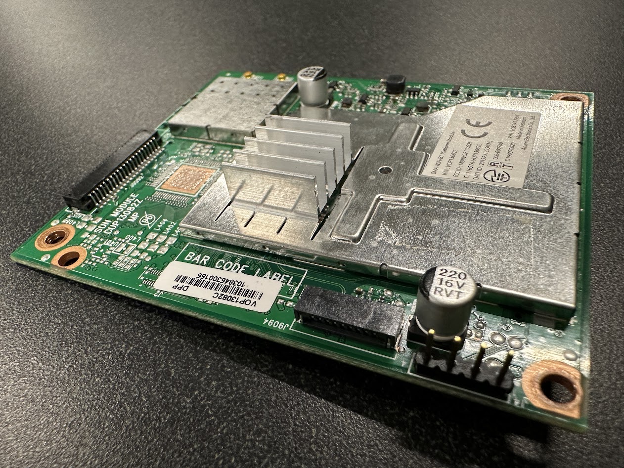 A close-up view of a printed circuit board with various electronic components, including a heatsink, capacitors, and a barcode label. The board is placed on a dark surface.