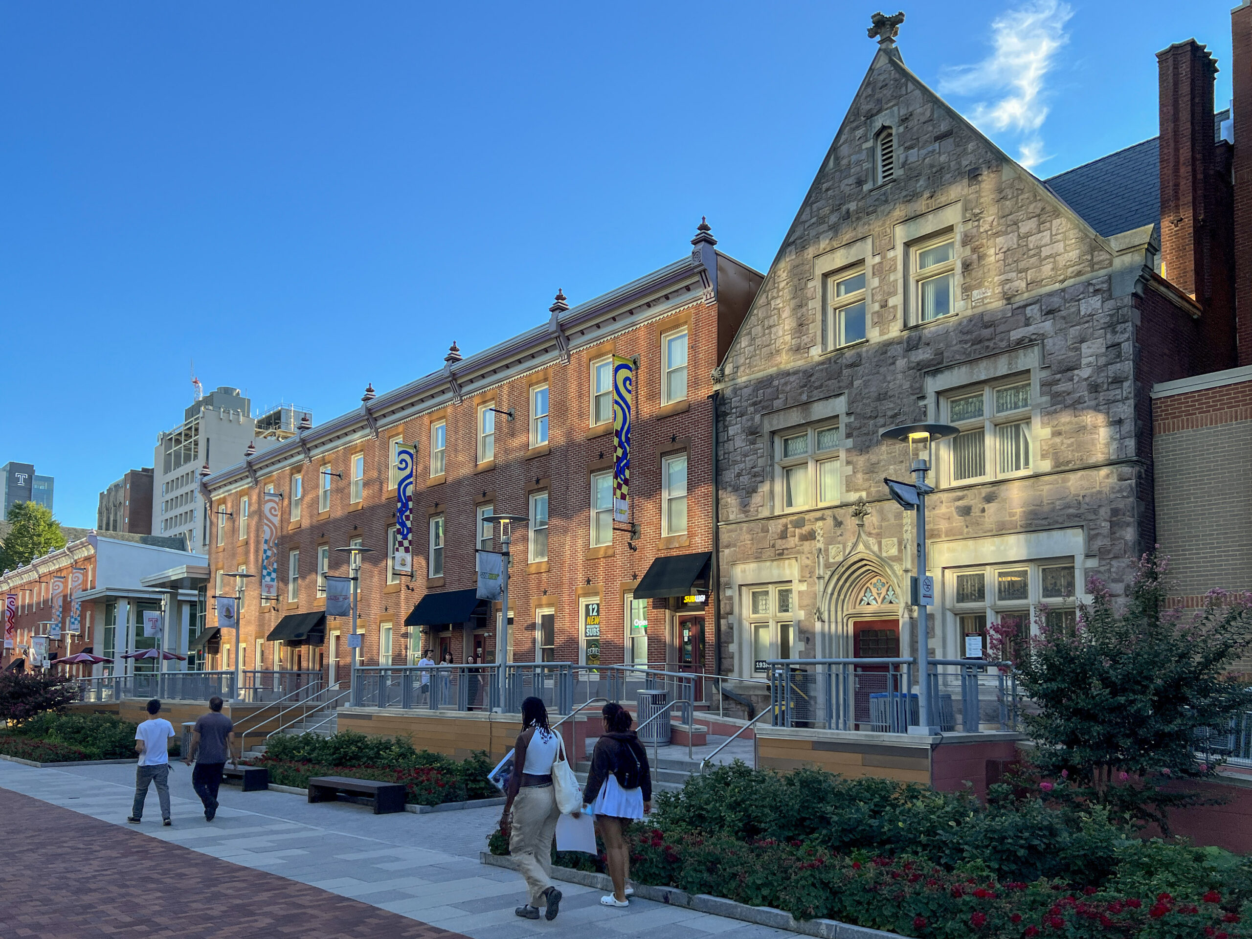 People walk along a brick path in front of historic buildings on a clear day. Some buildings feature red brick exteriors, while others are made of stone. Trees and benches line the path.