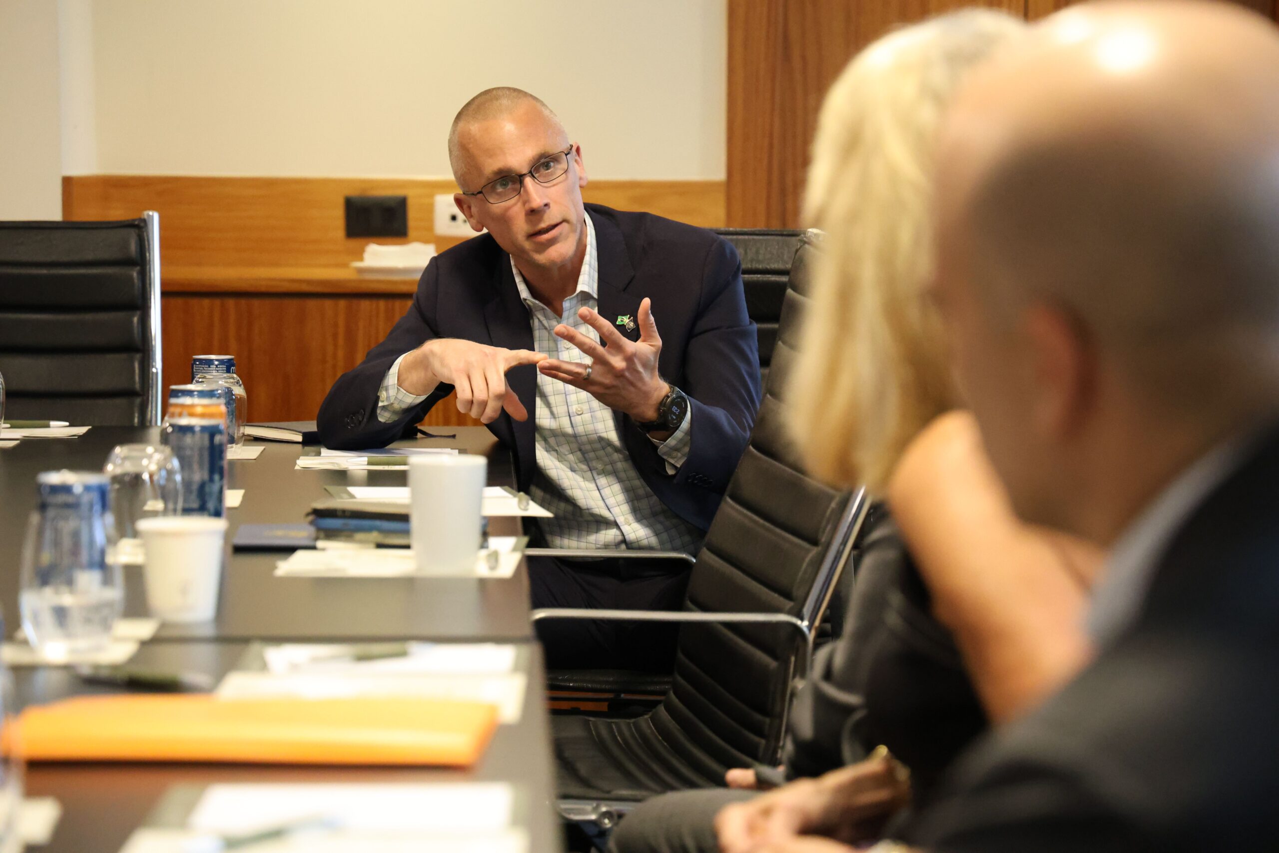 A man in a business suit is speaking and gesturing during a meeting, seated at a conference table with others.