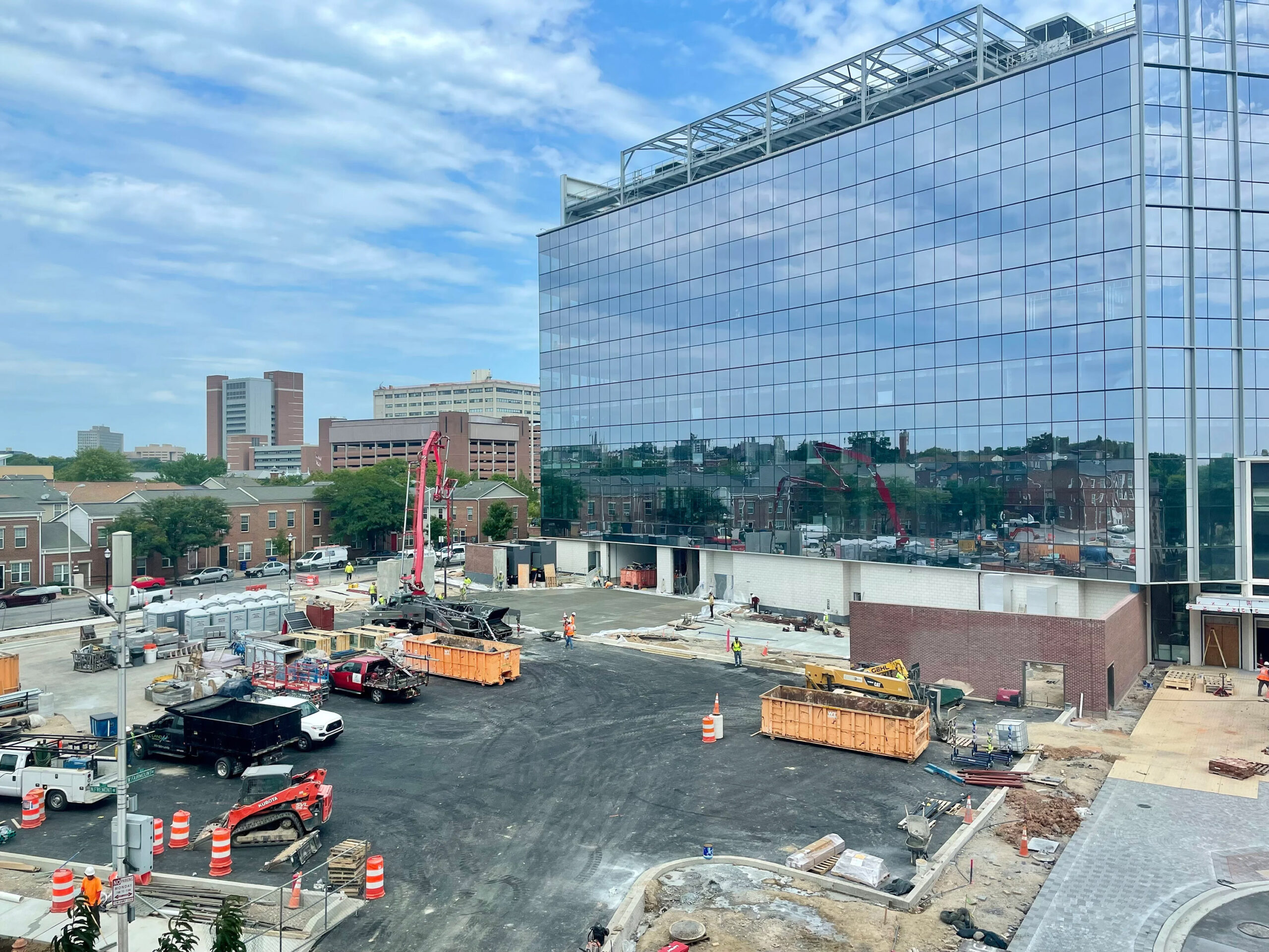 A construction site with heavy machinery and workers adjacent to a large glass office building. Orange traffic barrels and construction barriers are positioned throughout the area.