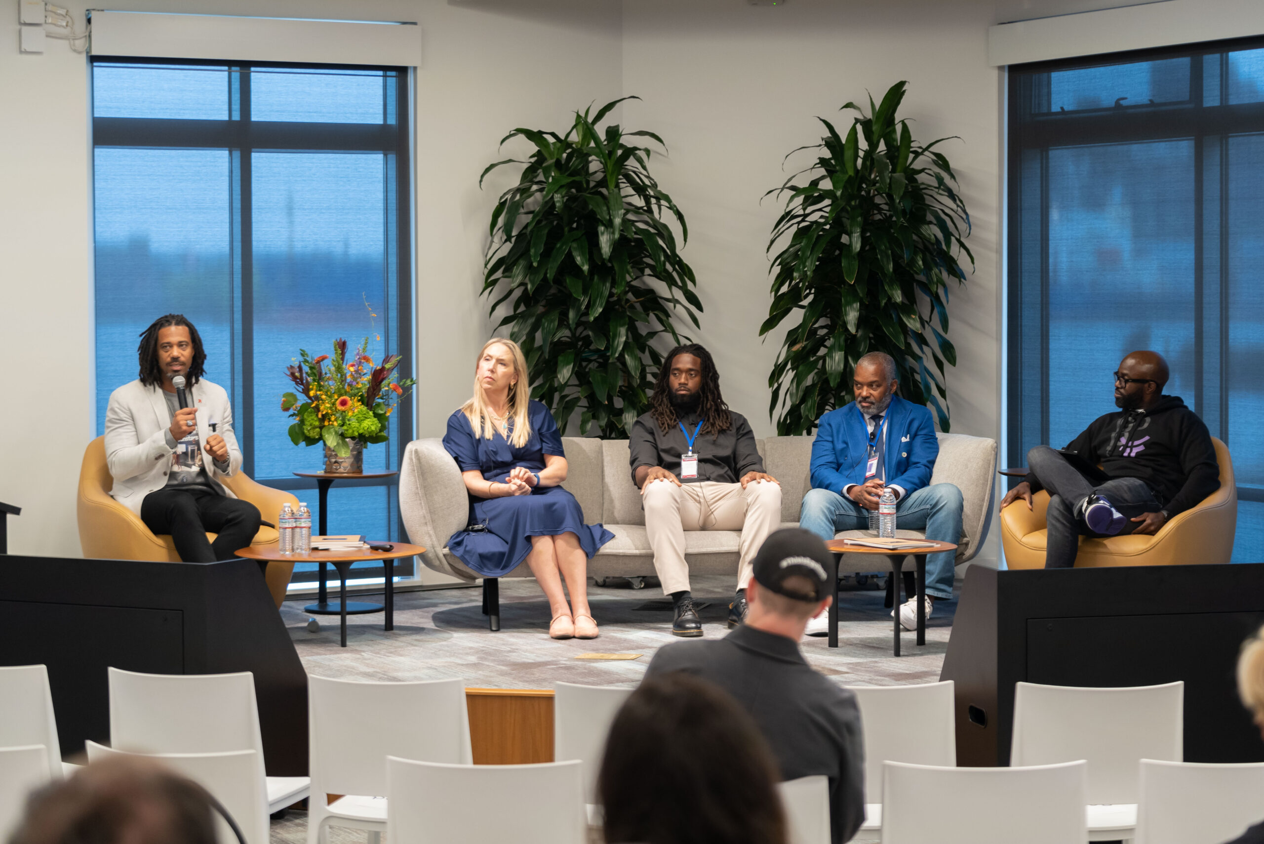 People on chairs and couches speak in front of green shrubs on grey stage