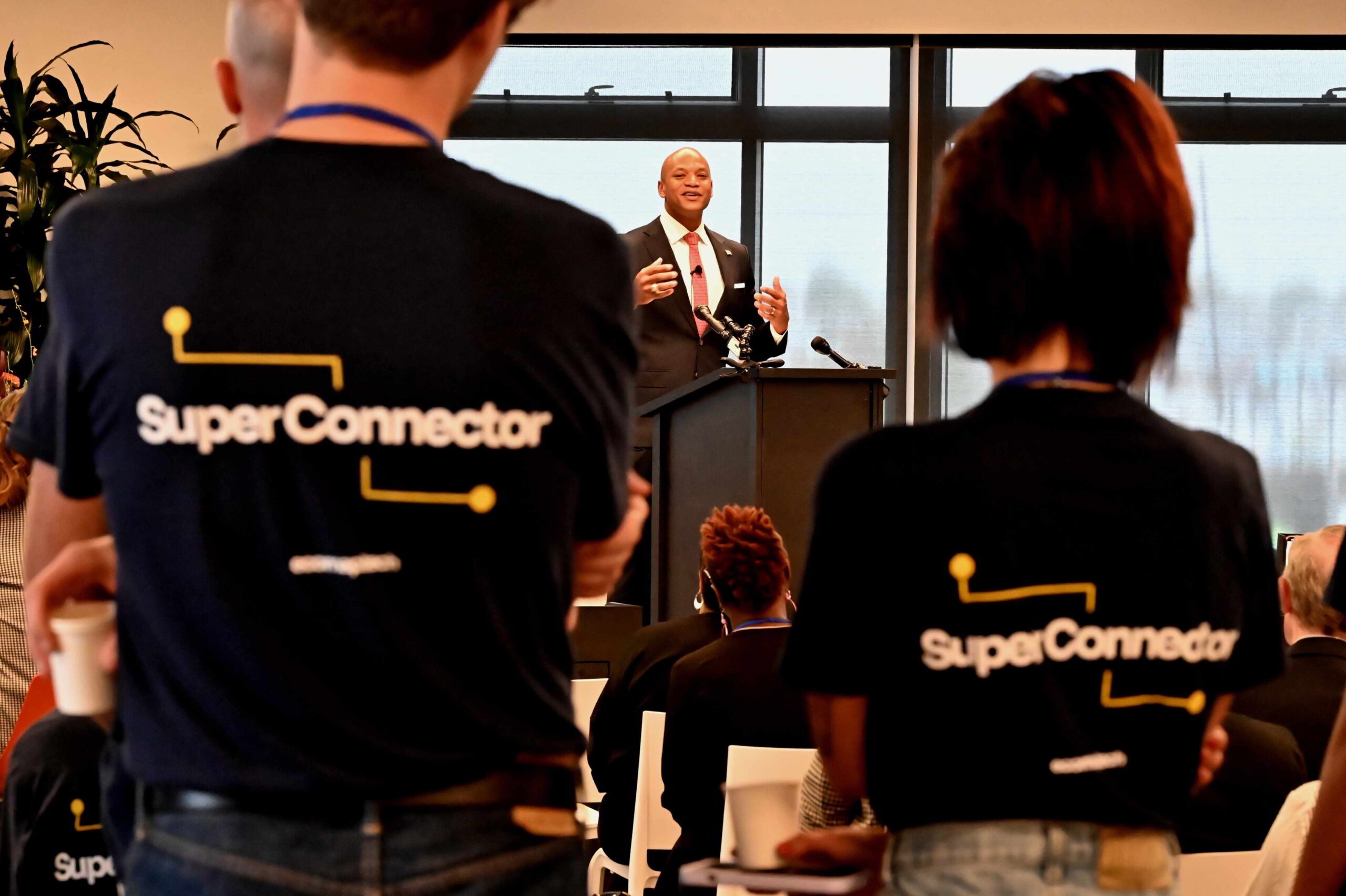 Gov. Wes Moore in charcoal suit on stage; people in shirts reading "SuperConnect" stand in foreground.