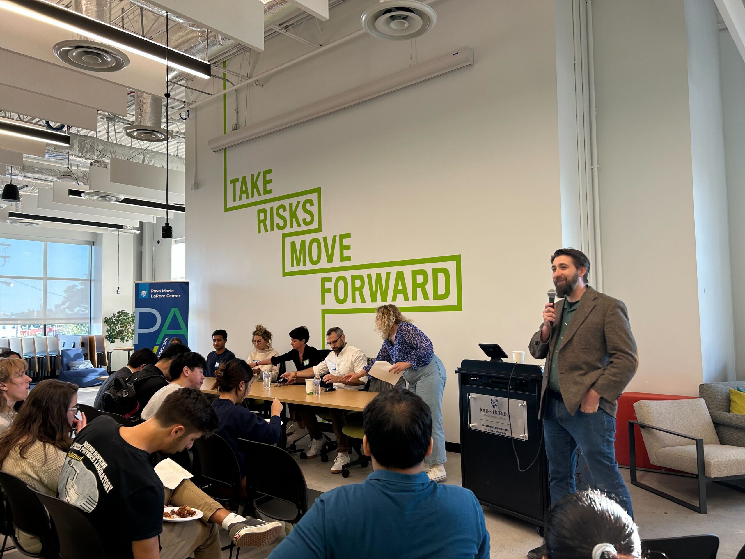 Man in brown blazer speaks into a microphone before an audience in black chairs, panelists at a brown table and a wall reading "TAKE RISKS MOVE FORWARD."