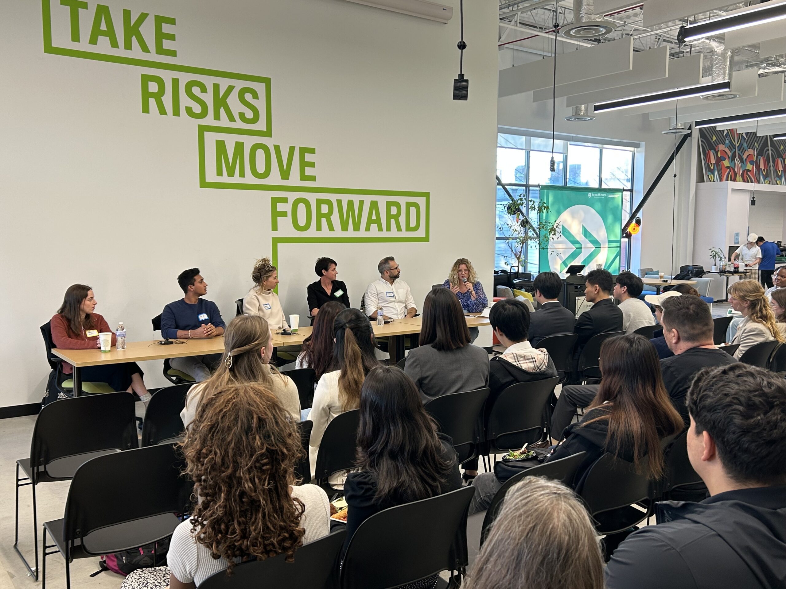 Six panelists speak to an audience in front of a wall reading "TAKE RISKS MOVE FORWARD" in neon green.