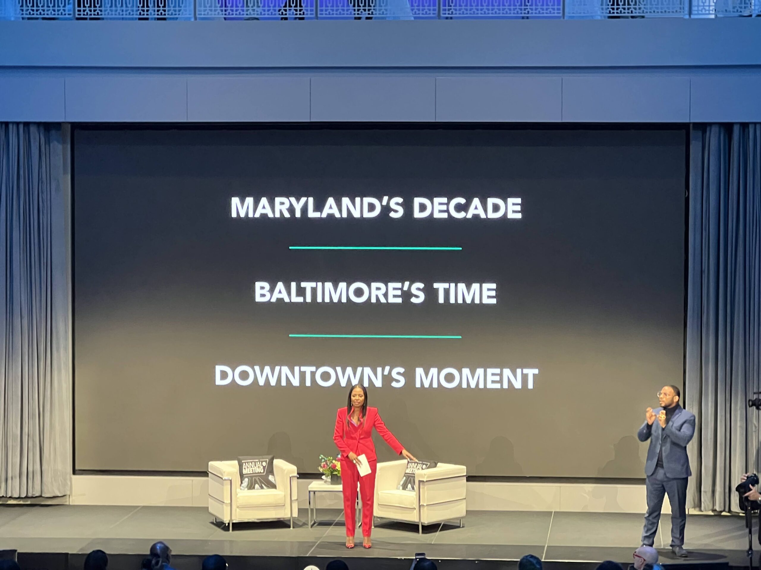 Woman in red suit speaks on stage with two cream chairs while man in navy suit  translates into ASL. A screen in the back reads MARYLAND'S DECADE, BALTIMORE'S TIME and DOWNTOWN'S MOMENT.