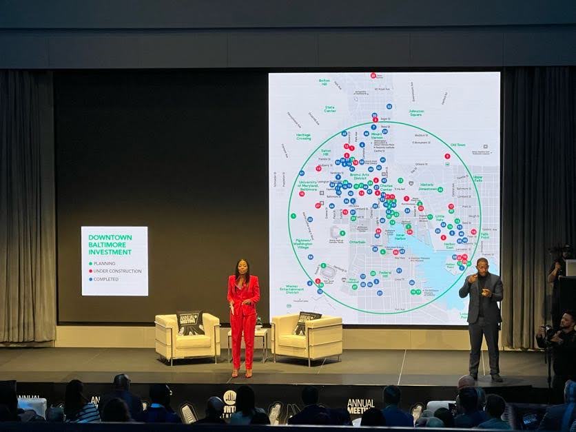 Woman in red suit and ASL signer in navy suit stand on stage in front of graphic showing Baltimore city with green, red and blue highlights of investment sites.