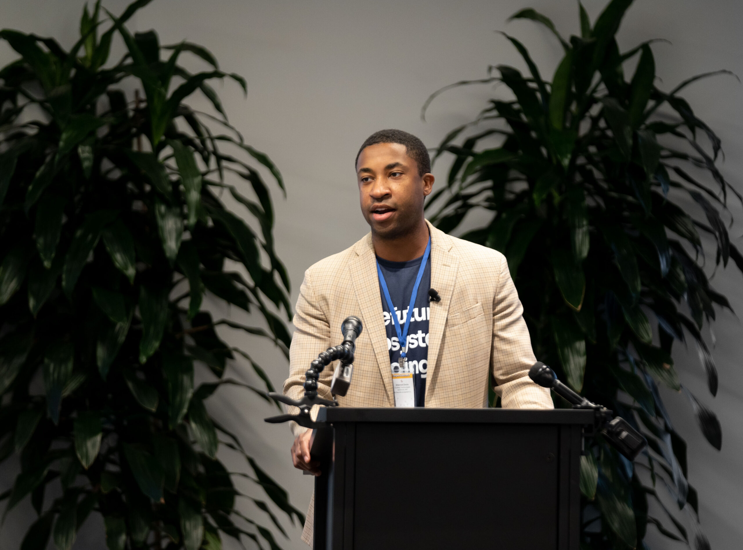 A Black man in a beige blazer and navy shirt speaks at a black podium in front of green plants.