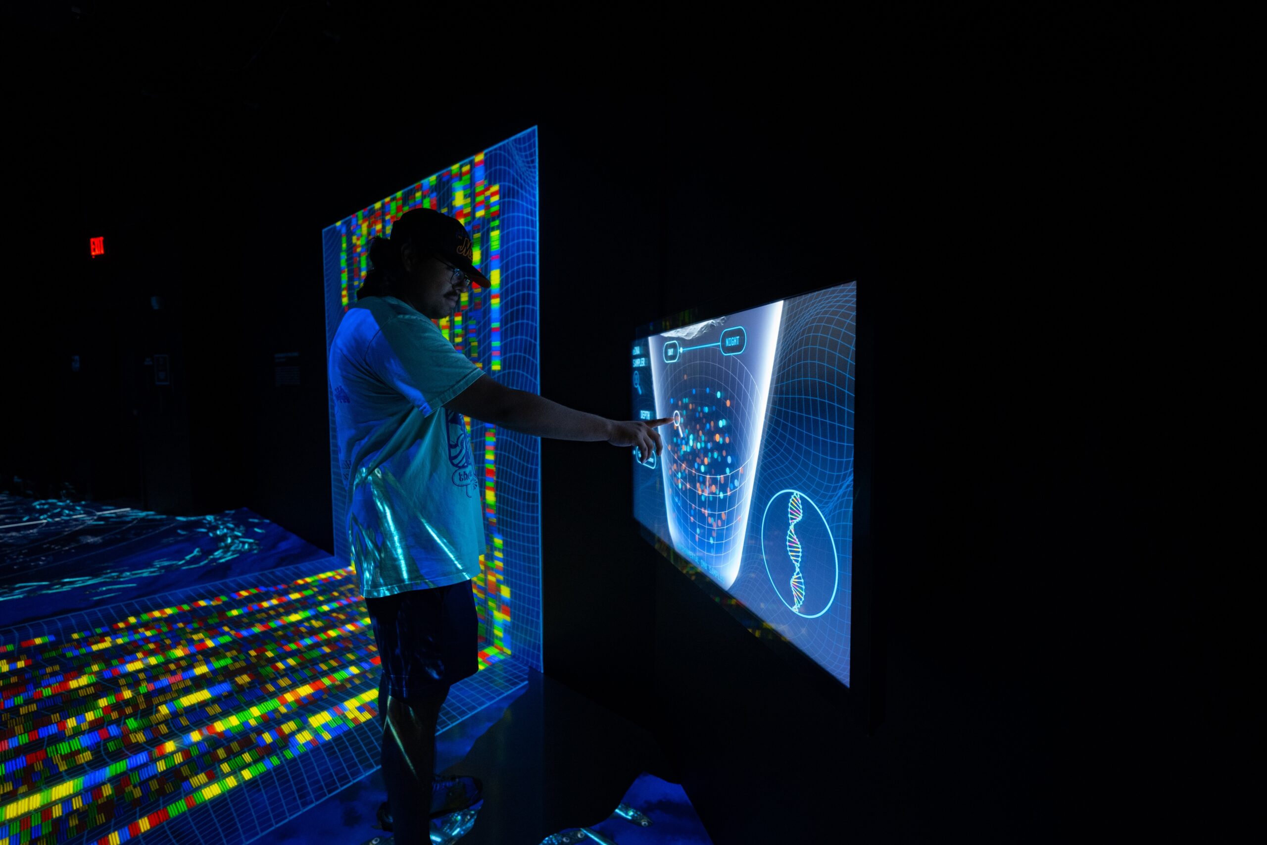 A person in a white shirt and black hat and jeans touches a rendering of genetic code in a multicolored immersive exhibit.