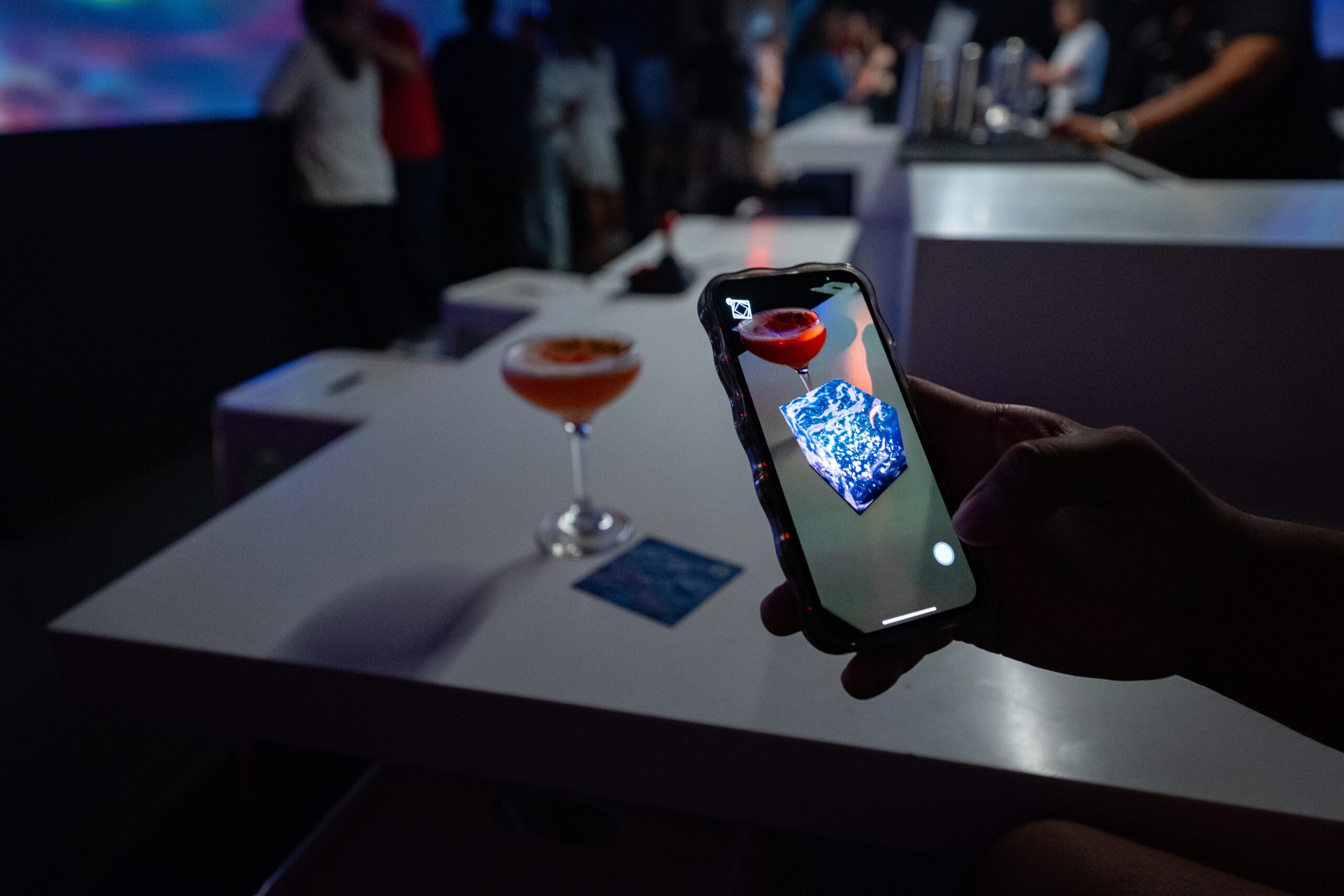 A person holds a mobile phone that shows an extended reality-rendered blue and white cube near a red cocktail on a bar.