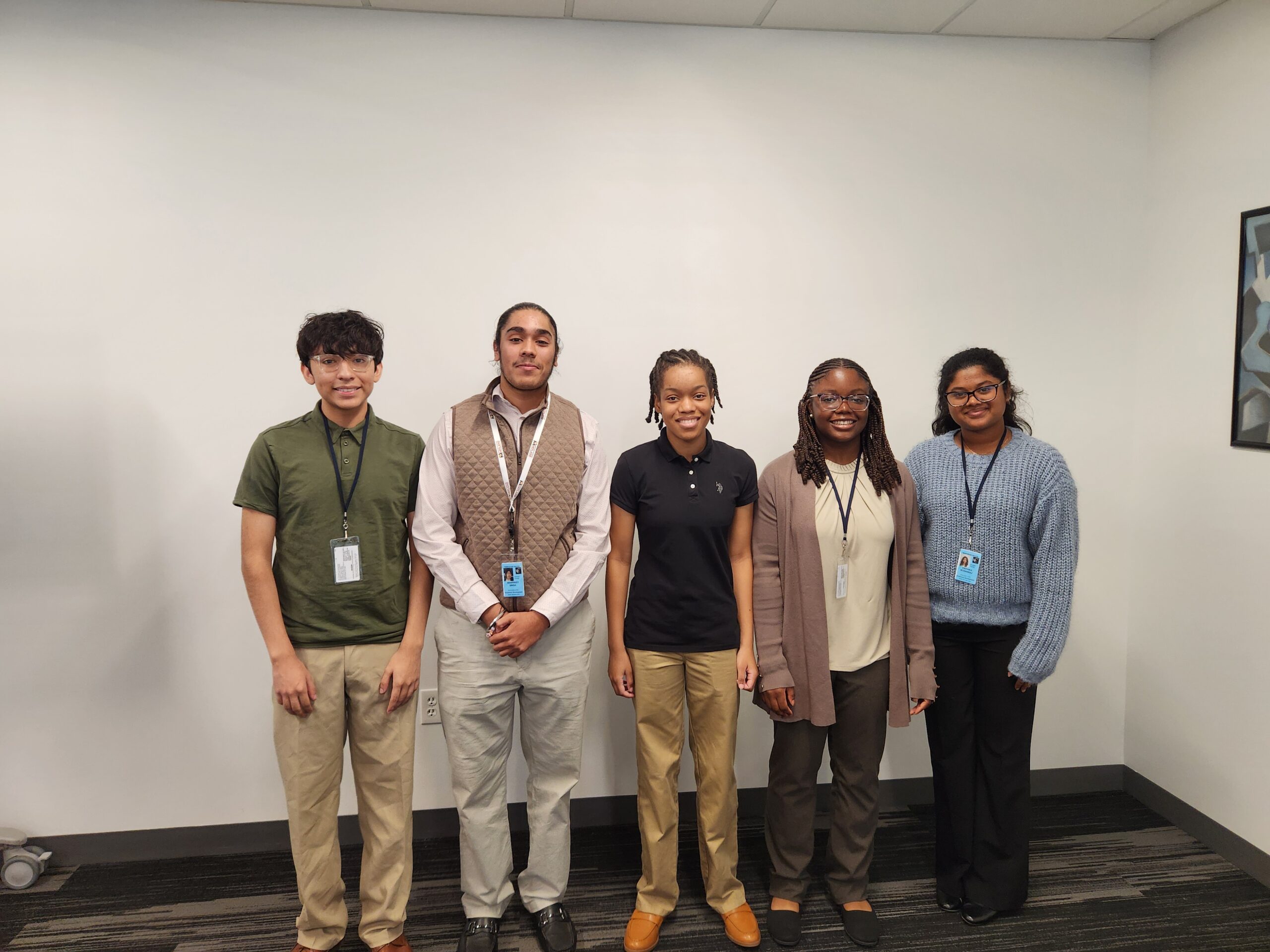 Five people standing in a row, wearing business casual attire, smiling at the camera in an office setting.